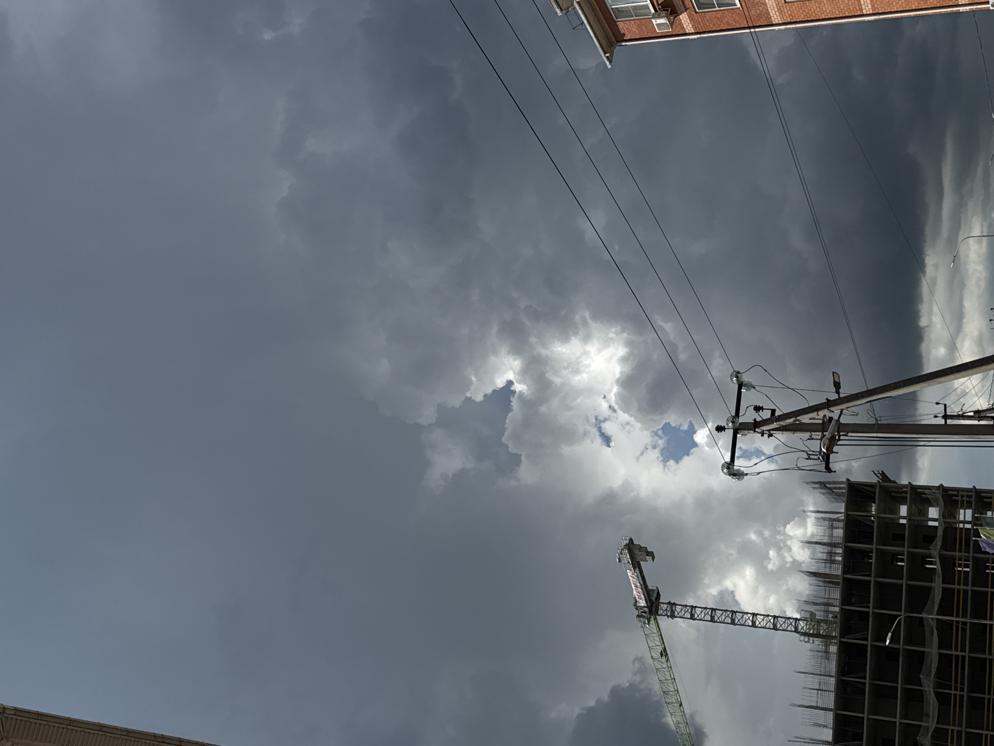The image captures a view looking upwards in what appears to be an urban environment. The sky is filled with heavy, dark clouds, suggesting an impending storm. There's a small patch of blue sky visible through a break in the clouds. Overhead power lines are visible, running diagonally across the frame. In the lower part of the frame, there's a construction site with a crane and the incomplete skeleton of a building. A brick building is partly visible at the top of the frame. The location is Unknown, Unknown.