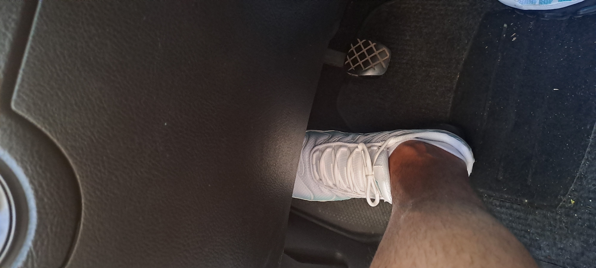 A close-up shot focuses on a person's right foot, clad in a white sneaker, resting on the floor mat of a vehicle. The sneaker has white laces tied in a bow and appears to be a running or athletic shoe. Adjacent to the foot is a diamond-patterned pedal, likely the clutch or brake pedal of a manual transmission car. The background consists of the dark, textured interior of the vehicle, possibly the dashboard or footwell area, with a faint indication of another foot or shoe in the lower right corner. The lighting suggests an indoor or shaded environment, possibly a car park.
