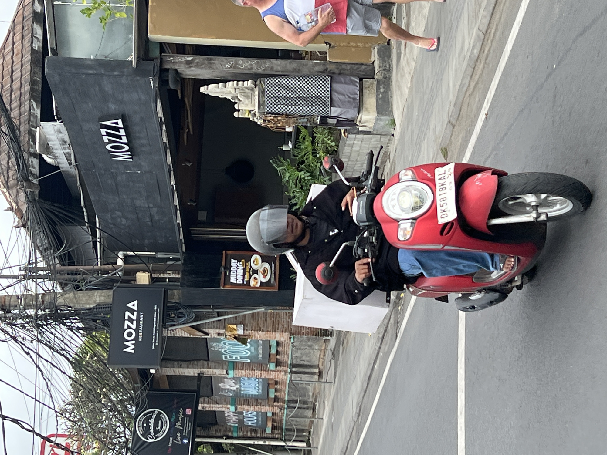 A red scooter is being ridden down a street in Denpasar, Indonesia. The scooter has a white license plate that reads "DK 5818 KAL". The rider is wearing a black jacket, blue jeans, and a helmet with a mirrored visor. In the background, there is a restaurant called "MOZZA". There are also several other signs and buildings visible. A man in a white, blue, and red tank top and grey shorts is walking on the sidewalk to the right of the scooter, holding two plastic bottles. The street appears to be paved with asphalt and there are power lines overhead.