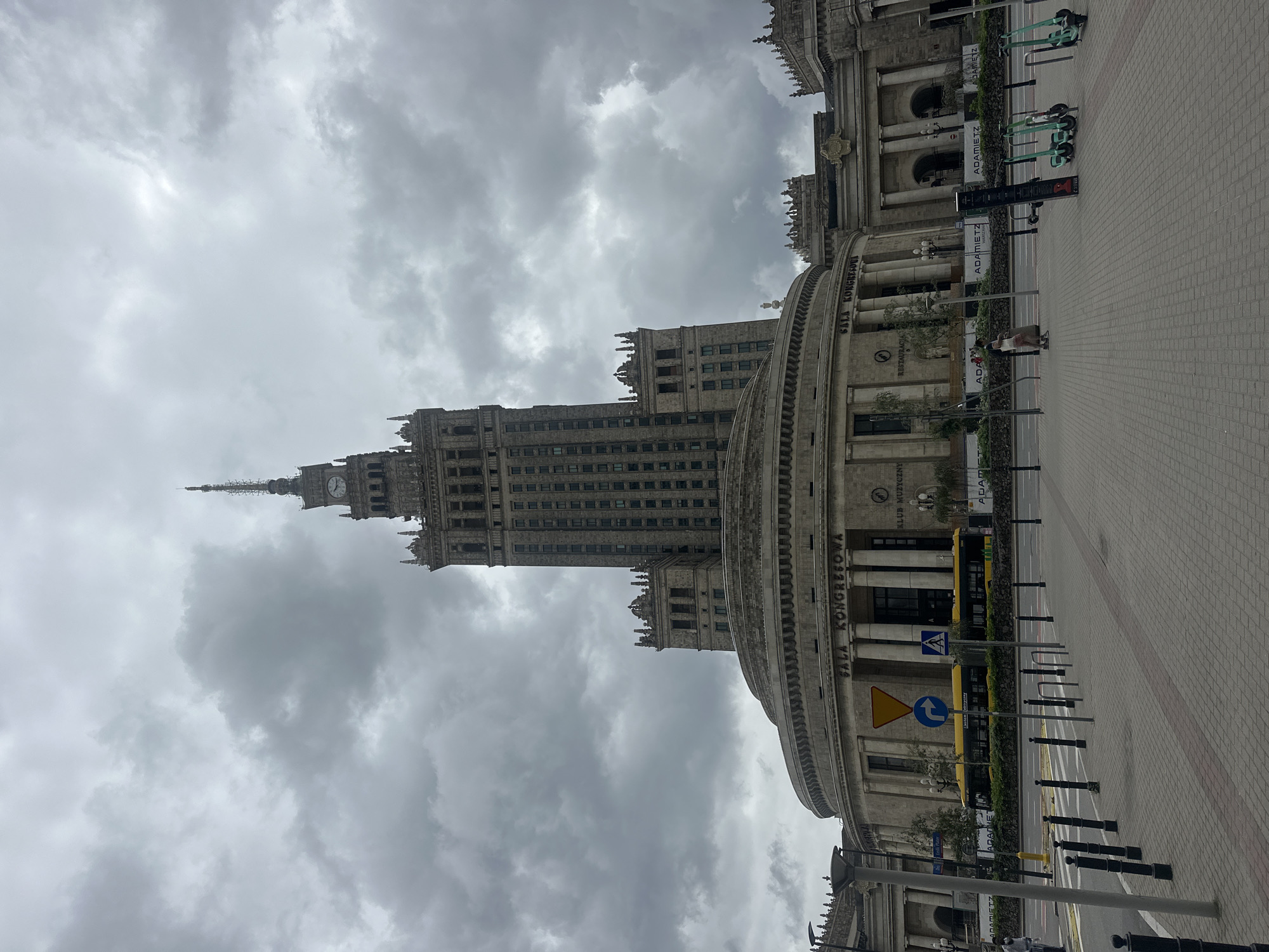 The image shows the Palace of Culture and Science in Warsaw, Poland, under a cloudy sky. The imposing building features a central tower with a clock and spire, flanked by lower, circular wings. The facade of the lower section is adorned with classical columns and the inscription "SALA KONGRESOWA" (Congress Hall) and "KLUB MUZYCZNY" (Music Club). In the foreground, a street is visible with a yellow bus, traffic signs, and bollards. Several electric scooters are parked on the sidewalk. Two individuals are walking on the sidewalk. The overall scene is urban and overcast.