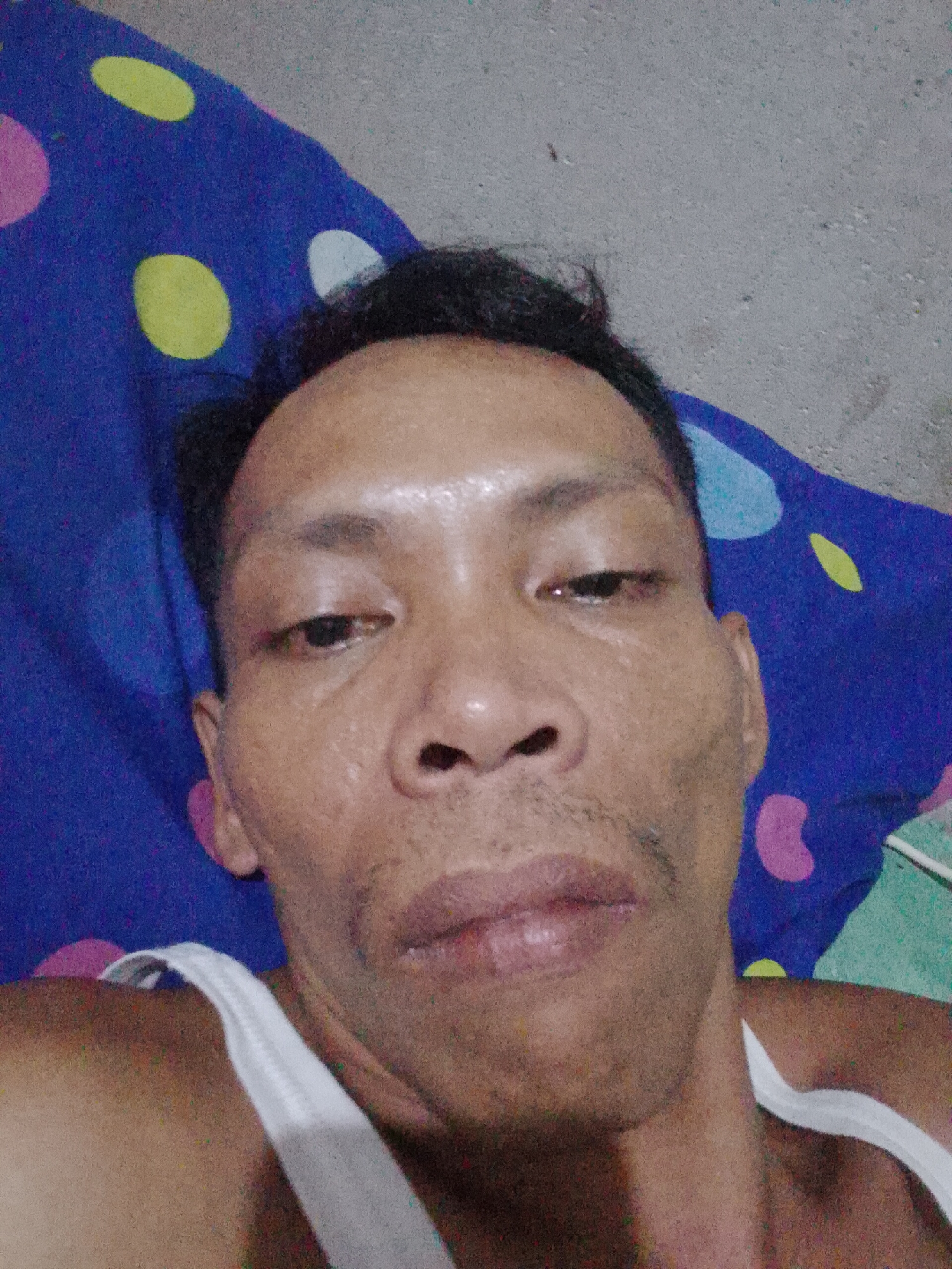 A close-up selfie shows a middle-aged man with short, dark hair and brown skin, lying down on a blue bedspread with yellow, pink, and white polka dots. He is wearing a white tank top. The background includes a concrete wall or floor. The location is Bukit Ketok, Indonesia.