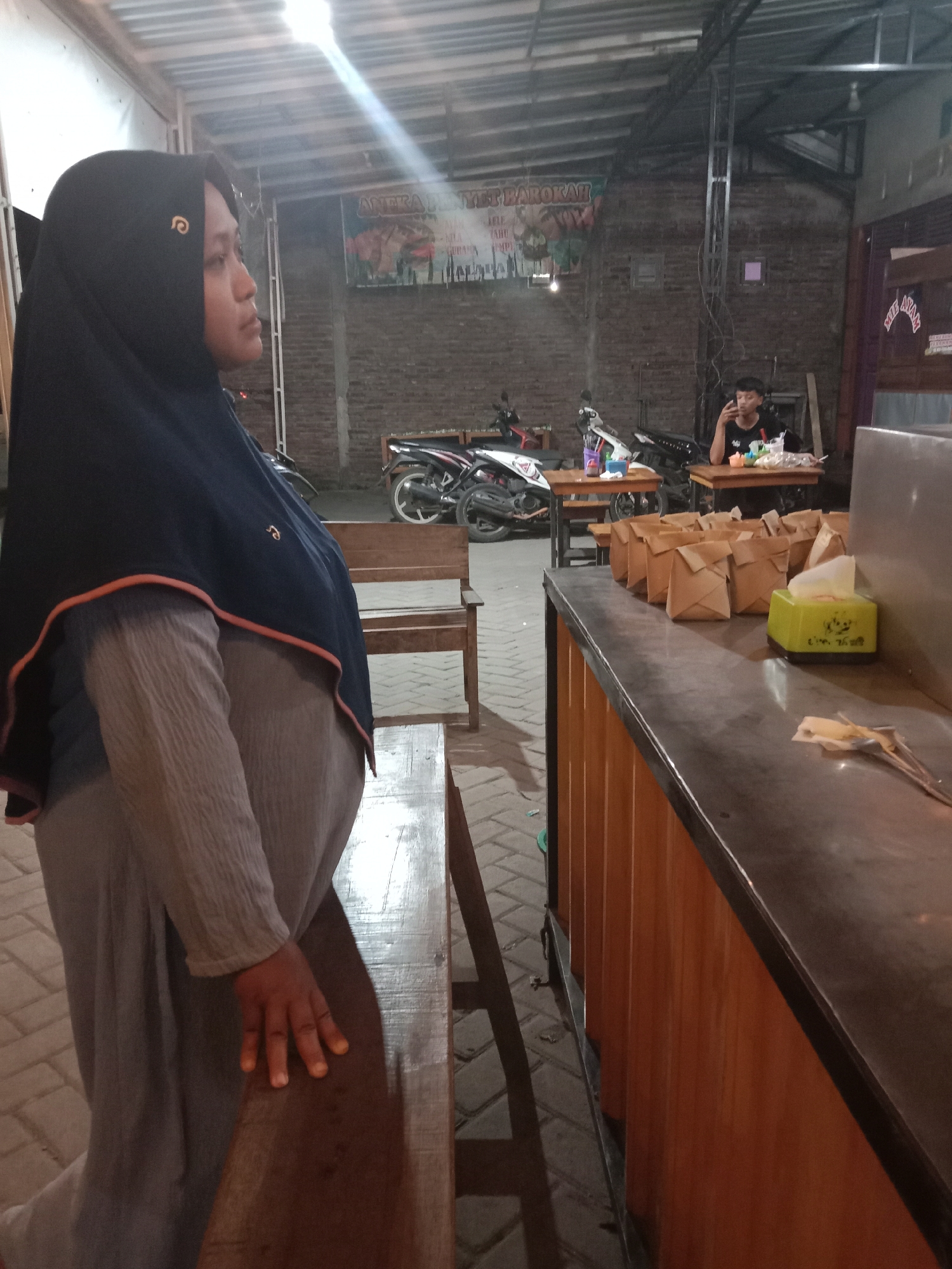 This image captures a nighttime scene in Cangkring, Indonesia, likely within a casual outdoor or semi-outdoor eatery. In the immediate foreground, a woman stands in profile, facing right, with her right hand resting on a dark wooden bench. She wears a dark blue hijab with an orange trim, adorned with a small gold-colored spiral pin on the left side of her head. Her attire also includes a light brown/grey textured long-sleeved top and a matching long skirt or trousers. She appears to be pregnant.
To the right, a dark-topped counter with vertical wooden paneling holds multiple light brown paper-wrapped food packages arranged in two rows. A bright green tissue box labeled "I'm the best" and a container of metal utensils are also present on the counter.
In the mid-ground, a young man is seated at a light-colored wooden table, facing slightly towards the camera while looking down at an object in his hand, potentially a mobile phone. Another wooden table and chairs are visible, along with two motorcycles parked between the tables and the background wall.
The background features exposed, light-brown brick walls. A banner on the left wall reads "ANEKA PENYET BAROKAH" with images of food items. Further right, a vertical sign partially displays "MIE AYAM". Overhead, a corrugated metal roof structure is visible, supported by metal beams. The scene is illuminated by artificial lighting, with a strong light source creating a visible beam in the upper center of the frame. The ground is paved with interlocking concrete bricks.