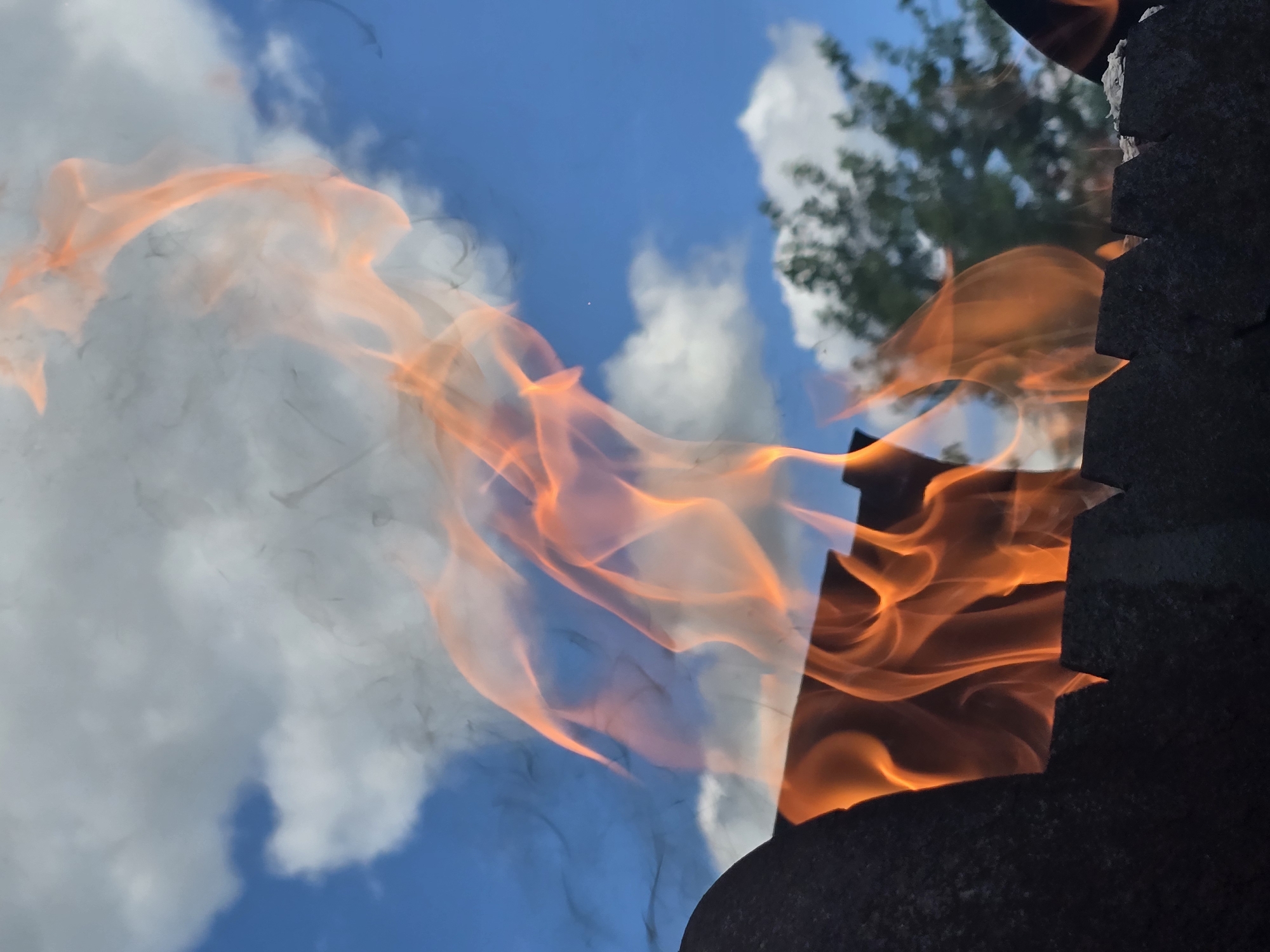 A close-up, low-angle shot captures flames rising from a charcoal grill against a bright blue sky dotted with white clouds. A dark, jagged rim of the grill is visible in the foreground, with hints of burning coals below the flames. Wisps of dark smoke curl upwards and to the left, mingling with the bright orange and yellow tongues of fire. In the background, the blurry green foliage of a tree can be seen against the sky, suggesting an outdoor setting, possibly a park or backyard in Ермолаево, Russia. The overall impression is one of a warm, summery day, with the fire being the central focus of the image.