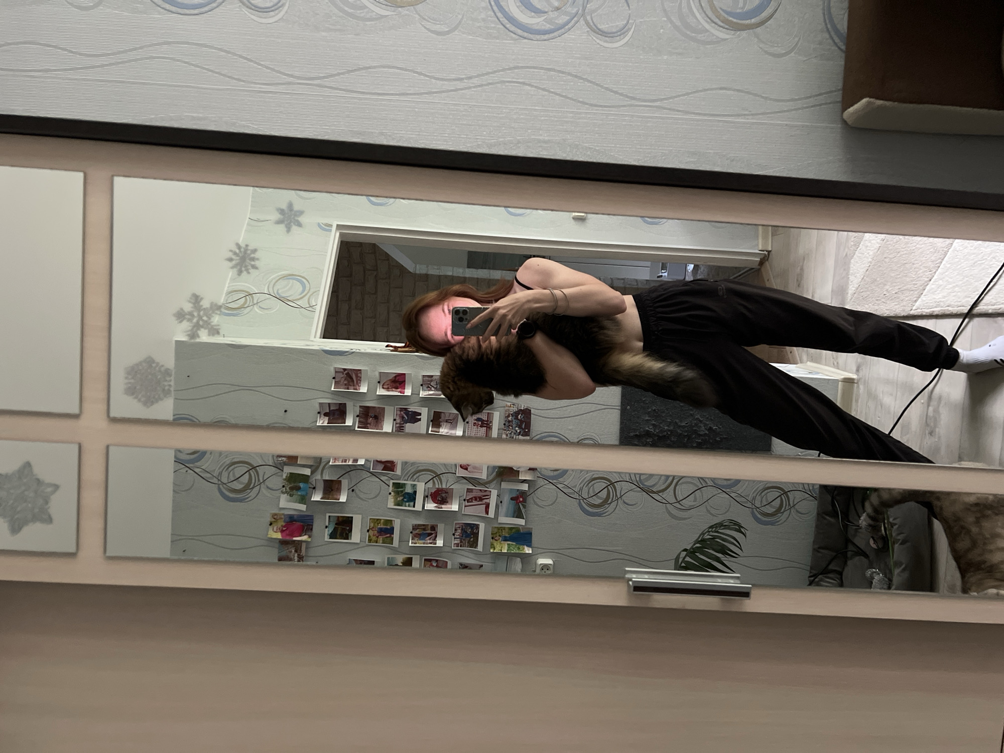 In the city of Chaikovsky, Russia, a young woman with reddish-brown hair is taking a selfie in a full-length mirror. She is wearing a black tank top and black sweatpants, holding a fluffy, multi-colored cat. The mirror is part of a closet with light beige doors. The background reflected in the mirror shows a wall with a light blue and grey patterned wallpaper decorated with snowflake decals. Another mirror, to the left of the main mirror, displays a collection of small printed photographs attached to the wall. Further to the right, a portion of a doorframe with a brick pattern is visible. The floor is covered with a light-colored rug. A cat bed is visible in the reflection of the left mirror. The overall scene suggests an indoor residential setting.