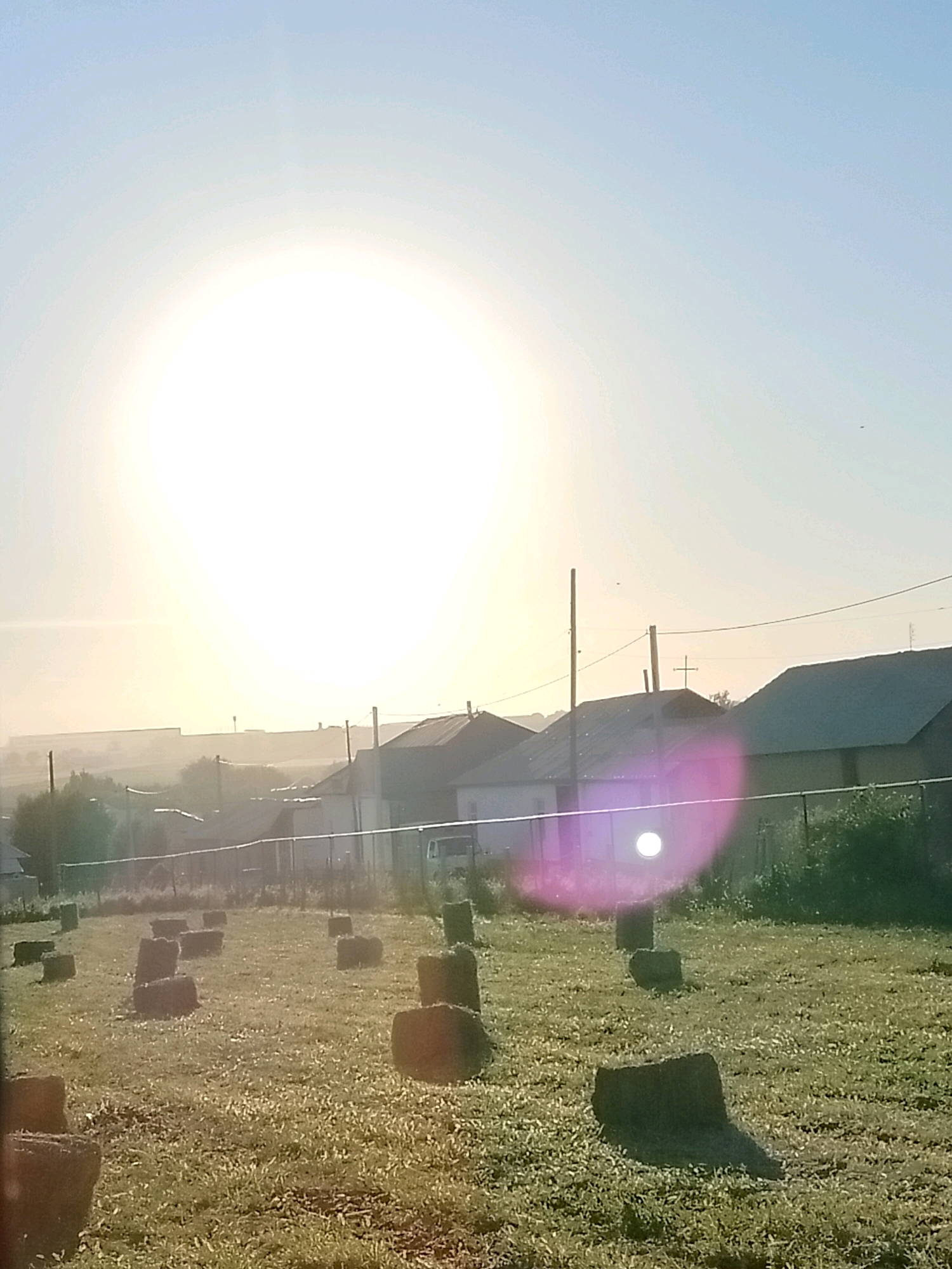 In the city of Көлкент, Kazakhstan, an outdoor scene unfolds under a bright, hazy sky, dominated by the sun's intense glare. The foreground of the image features a grassy field scattered with numerous rectangular bales of hay. Beyond the field, a line of low-rise buildings with dark roofs is visible, separated by a chain-link fence. A utility pole stands to the left of the frame, and electrical wires are strung across the sky. In the distance, more structures and trees are faintly discernible through the atmospheric haze. A distinct pinkish lens flare is present in the lower right quadrant of the image, partially obscuring some of the buildings. No people are visible in the frame.
