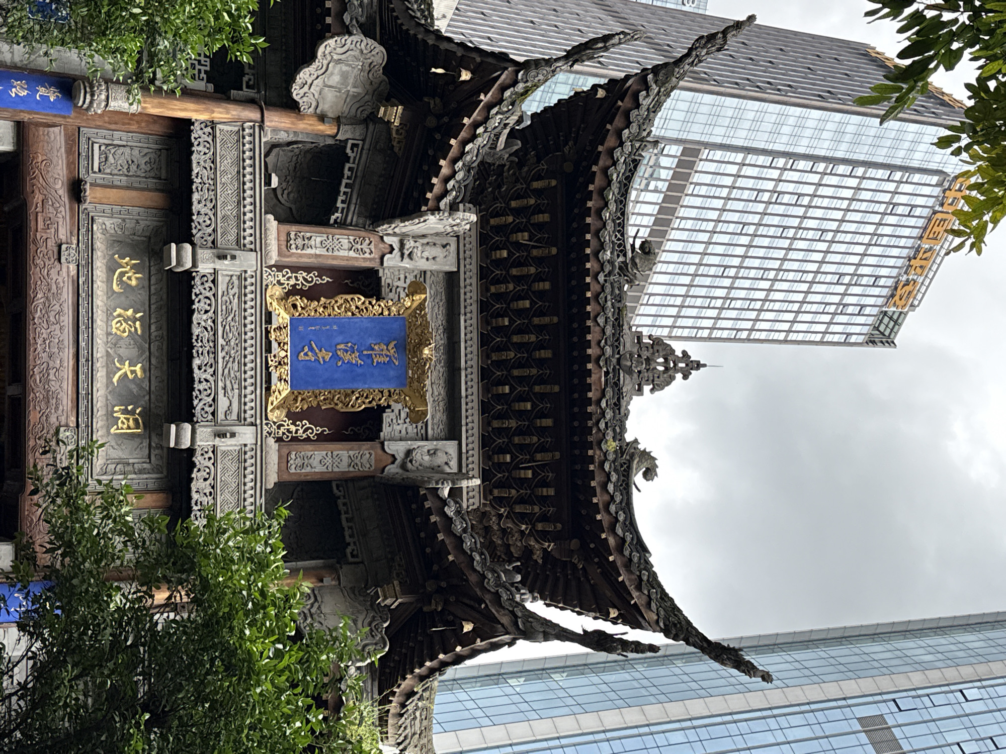 An intricately detailed traditional Chinese architectural structure, possibly a gate or temple entrance, stands prominently in the foreground, contrasted sharply against a modern high-rise building under an overcast sky. The traditional building features a multi-tiered, ornate roof with upturned eaves and decorative elements that appear to be carved stone or wood. The roof's edges are adorned with mythological figures or intricate patterns.
Below the roofline, a large, rectangular blue plaque with gold Chinese characters reads "通远门" (Tōngyuǎnmén). To the left, a smaller, partially visible blue plaque also bears Chinese characters, likely related to the main sign. Gold Chinese characters, "大福地" (Dàfúdì), are visible vertically on a wooden beam or door frame of the traditional structure. The traditional building itself is rich in detail, with complex carvings on its wooden pillars, beams, and decorative panels. Green foliage from a tree frames parts of the traditional structure, adding a touch of nature.
Behind this historic edifice, a towering modern skyscraper dominates the background. Its facade is composed almost entirely of reflective glass panels, creating a grid pattern that mirrors the light. On one side of the modern building, large golden Chinese characters are visible, reading "融创" (Rongchuang), indicating a company name, likely a real estate developer. The setting appears to be an urban area where ancient cultural heritage coexists with contemporary development. The time of day is daylight, with the muted sky suggesting an overcast day. No people are visible in the scene.
