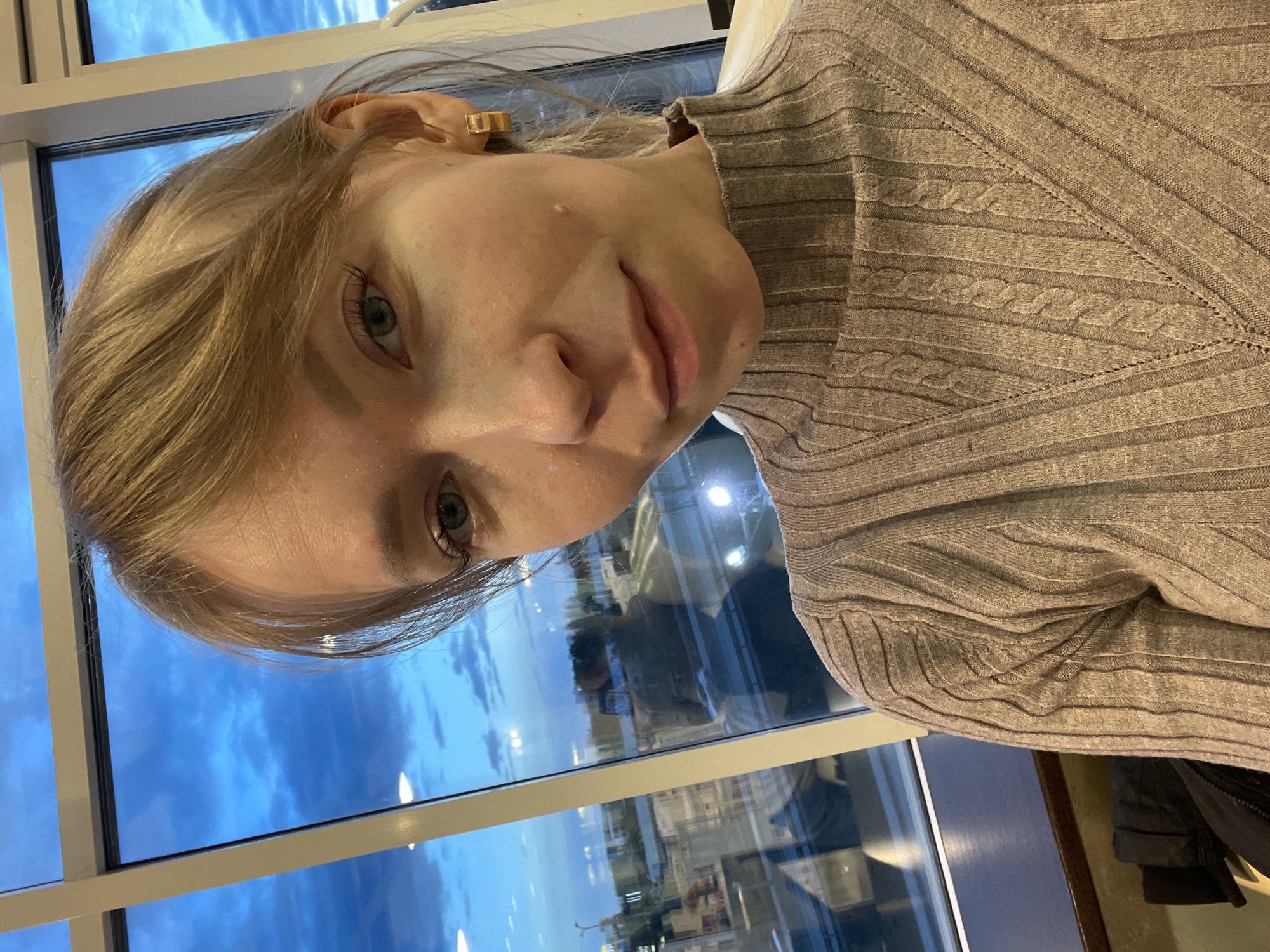 A fair-skinned person with blonde hair and wearing a brown cable-knit turtleneck sweater is indoors, positioned near a large window that is reflecting the exterior. The window appears to be multi-paned. Reflected in the glass, a city scene is visible with buildings and sky present. Based on the reflection of urban structures and the sky, the location is suggested to be Perm, Russia. The person is looking towards the camera. Their earring is a simple gold geometric design. The lighting suggests it may be dusk or dawn, with a blueish hue to the sky.
