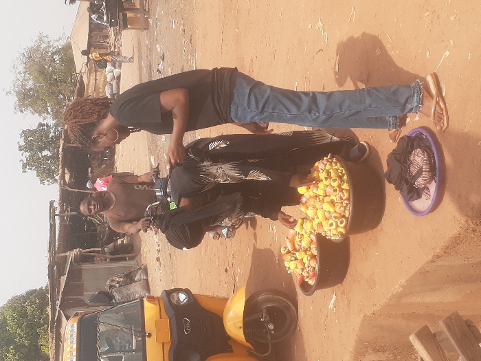 The scene depicts a bustling outdoor market area in Gwadanye, Nigeria, characterized by reddish-brown dirt ground and makeshift structures. In the foreground, two black plastic basins filled with yellow and red cashew apples and nuts are positioned on the ground. A woman wearing a black top and blue jeans, with dreadlocks and large hoop earrings, stands to the right, looking down towards the cashews. Another woman, dressed in a black abaya and headscarf, is bent over the basins, appearing to interact with the produce. Behind them, a man in a brown t-shirt sits astride a black motorcycle, gazing directly forward. To the far left, the front section of a yellow three-wheeled auto-rickshaw ("Keke Napep") is visible. In the immediate foreground, a purple plastic tray rests on the ground, holding dark fabric and a small patterned bag. The background features various crude market stalls constructed from wood and corrugated metal, with sacks of goods visible, and sparse trees under a hazy sky.