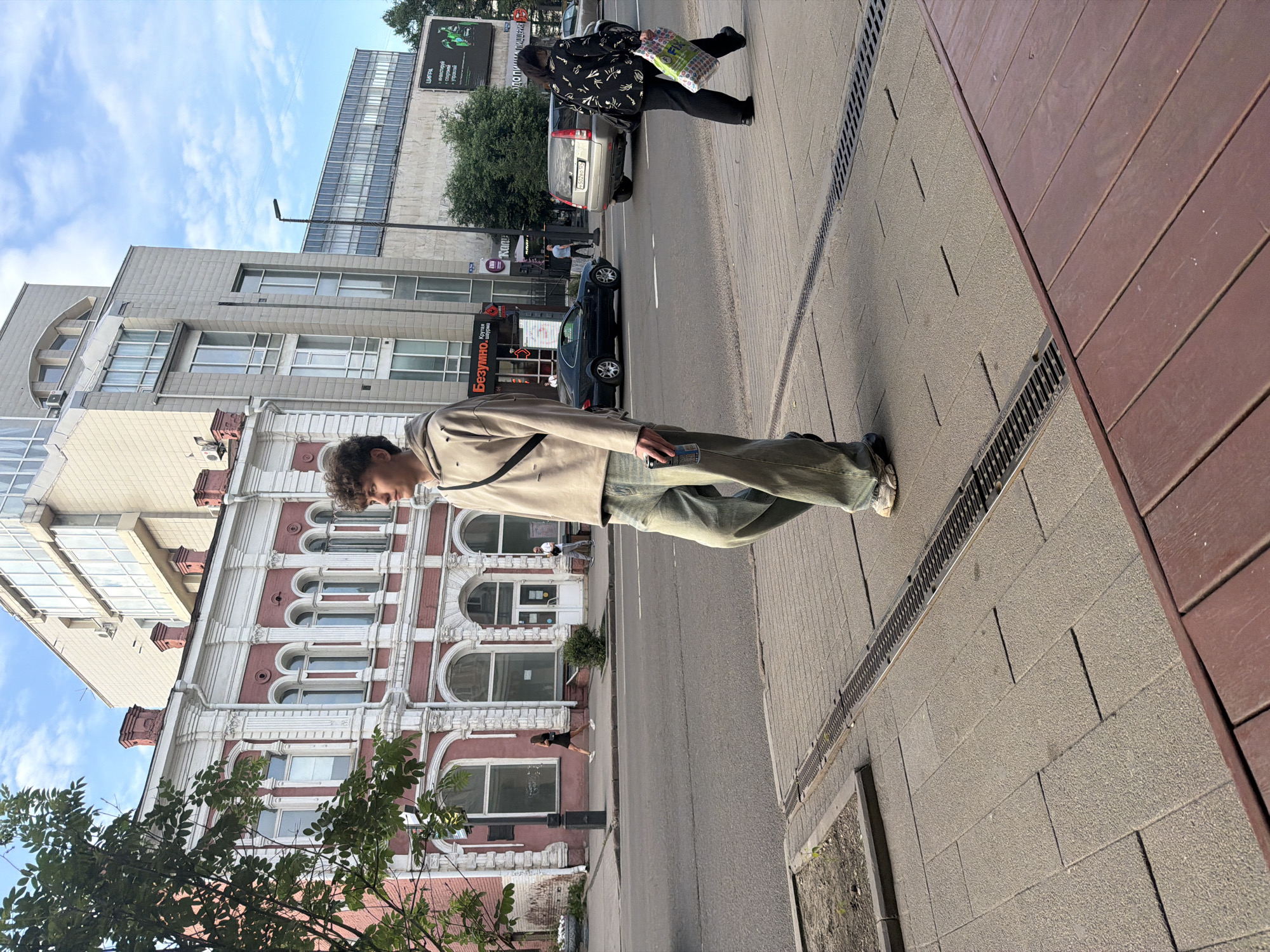 The image depicts a street scene in Krasnoyarsk, Russia. In the foreground, a young man with curly brown hair is walking on a sidewalk, holding a beverage can. He is wearing a beige hoodie with ripped details and light-wash, baggy jeans. A dark crossbody bag is slung over his shoulder.
Behind him, a street with a few cars is visible. A silver SUV and a black sedan are parked or moving on the road. A woman in a black jacket with a white pattern is walking on the sidewalk on the right side of the image, carrying a colorful shopping bag.
The background features several buildings. On the left, there is an older, pink building with white ornate decorations and arched windows. Adjacent to it is a modern, grey building with a glass facade. Further down the street, more buildings and trees can be seen. The sky is partly cloudy. The image appears to have been taken during the daytime, likely in the afternoon, given the lighting and shadows.
