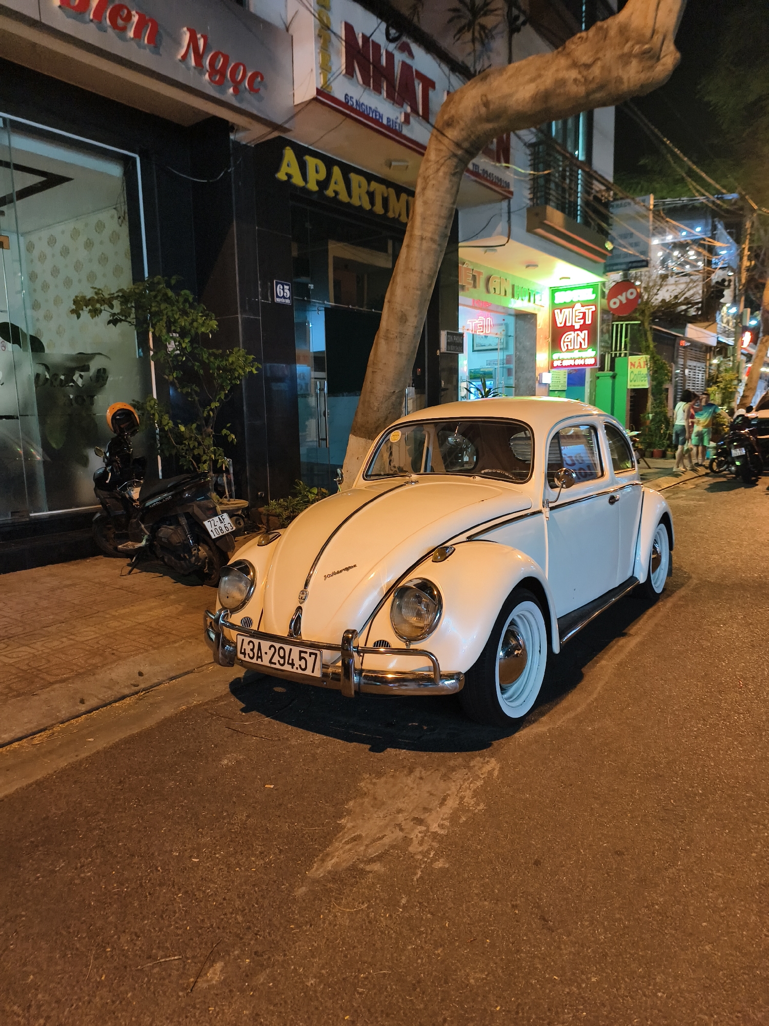A white Volkswagen Beetle is parked on the street in front of a building with a sign that reads "APARTM." A tree trunk obscures part of the building's facade. To the left, a parked motorcycle with a yellow helmet visible. In the background, signs for "Việt Ân" and "NA" Coffee are visible. People are standing and conversing in the distance. Street lights illuminate the scene. The car's license plate reads "43A-294.57." The buildings in the background include the address "65 NGUYEN BIEU."