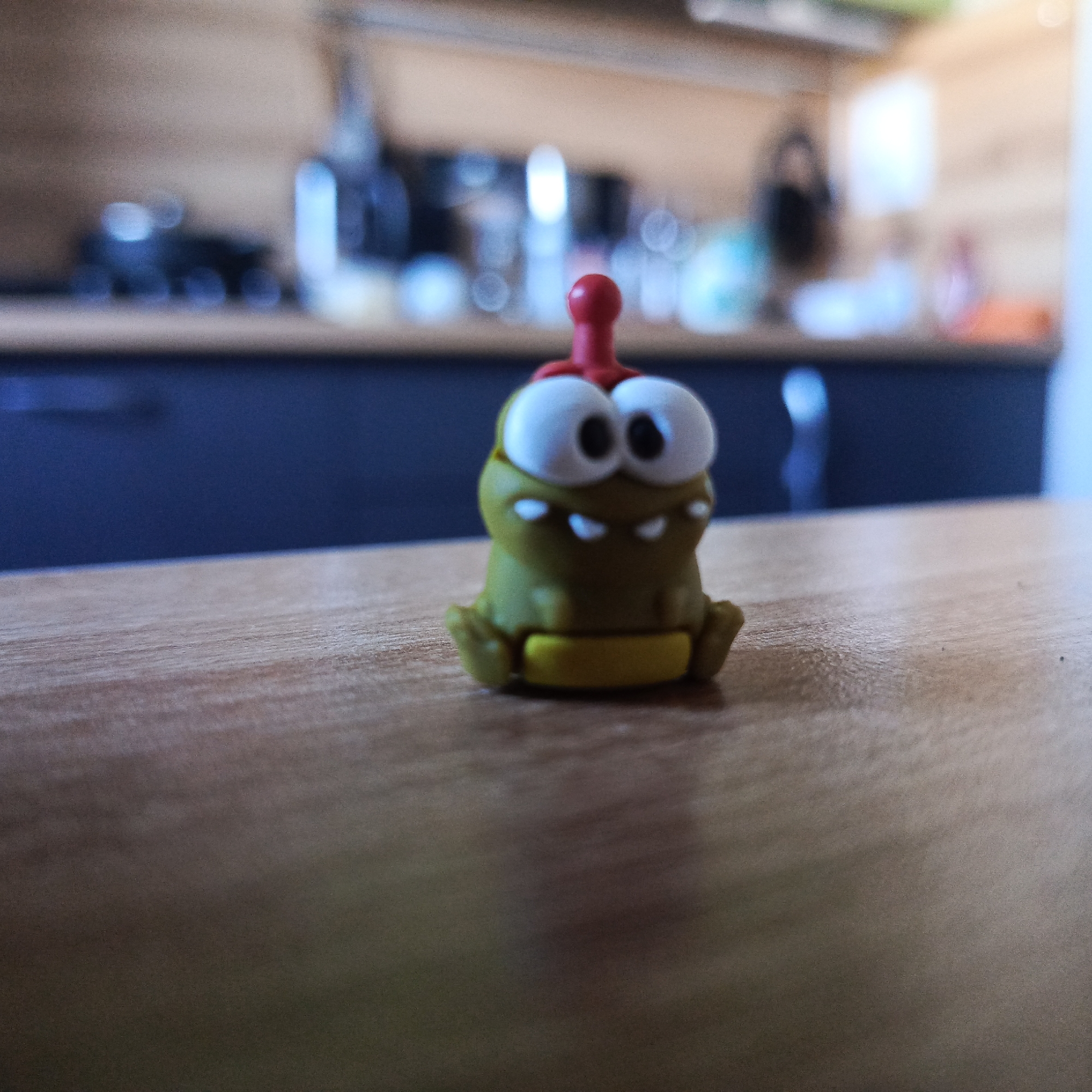 A close-up, shallow depth-of-field photograph captures a small, olive-green toy figure, resembling Om Nom from the "Cut the Rope" game, sitting on a light brown wooden table or countertop. The figure has large, white, round eyes with black pupils and a small, slightly open mouth showing three white teeth on its upper jaw. A red, thin, antennae-like protrusion extends upwards from its head. The toy's base is yellow, and it appears to be in a seated position with small, squat arms and legs.
The background is significantly out of focus, creating a bokeh effect with various blurred light sources and indistinct shapes. Behind the table, a dark blue or black surface, possibly a cabinet or lower wall, is visible. Above this, a lighter brown surface extends across the upper part of the background, likely another cabinet or wall section. The blurry shapes suggest common kitchen or indoor items, but they are too indistinct to identify precisely. No specific activity is occurring; the toy is simply posed. There is no visible text in the image. The image was taken in Марковский, Russia.