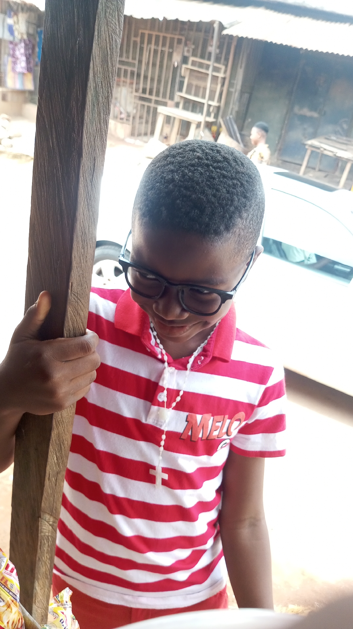 A young boy stands in front of a building in Nkpor, Nigeria. He is wearing a red and white striped shirt, glasses, and a necklace with a cross pendant. He is holding onto a wooden post with his left hand. In the background, there is a building with a metal gate and tables. A man is visible in the background behind a parked car.
