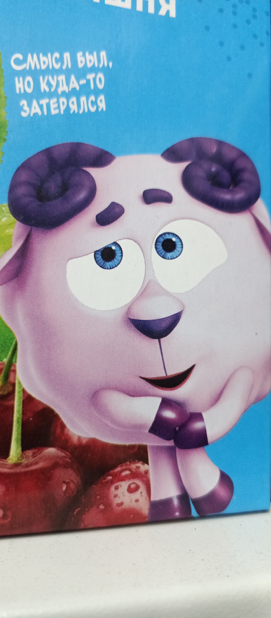 The image shows a close-up of a cartoon character, a purple sheep with large blue eyes and curly horns, on a bright blue background. The sheep is in the foreground, its head and upper body visible. Below the sheep, a portion of a pile of red cherries is visible. White text is present in the upper left corner of the image. The text appears to be in Cyrillic script. The overall image is a still from what appears to be a children's cartoon or promotional material. The location is not discernible from the image.