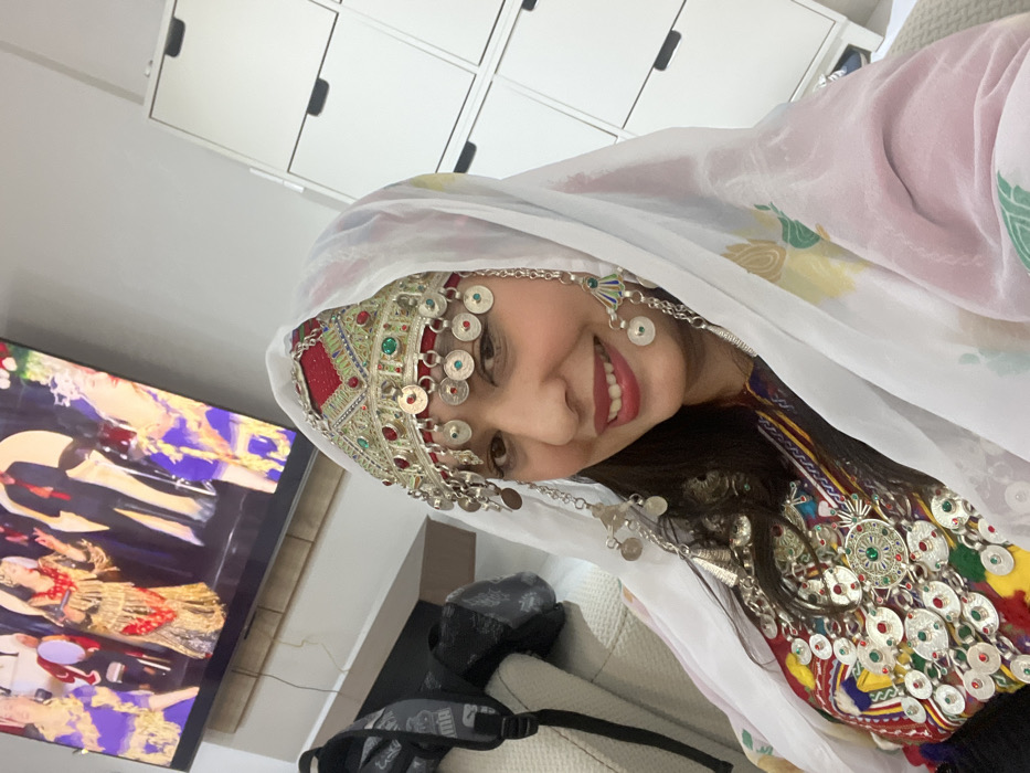 A woman wearing traditional clothing smiles at the camera. She wears a white headscarf, ornate headpiece with dangling silver coins, and a colorful necklace. In the background, a television displays a performance with dancers wearing elaborate costumes. A white cabinet with black handles is visible above. A grey bag with black straps rests on a light-colored surface. The scene is brightly lit, creating a cheerful and festive atmosphere.