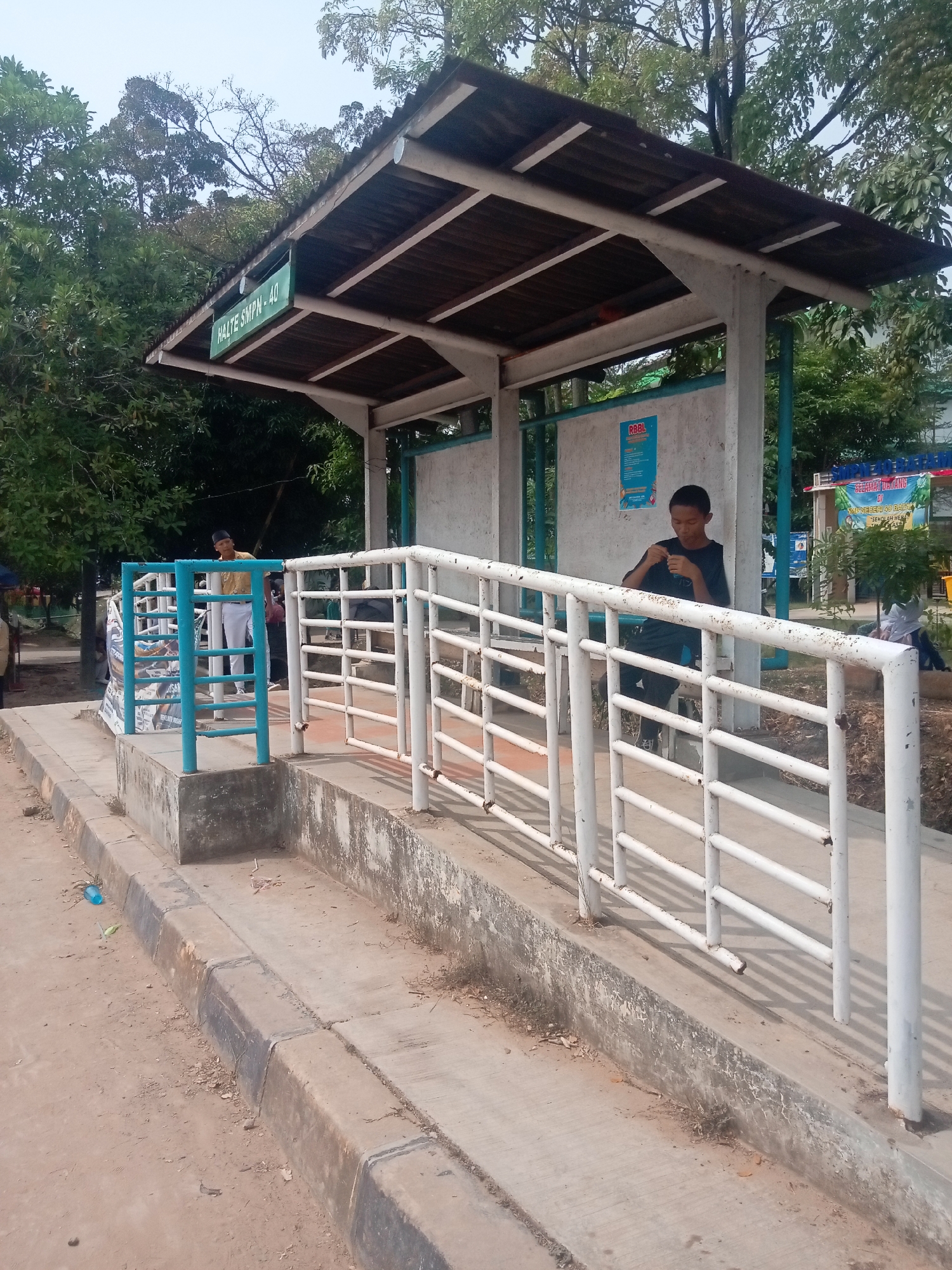 The image shows a bus stop shelter in Batam, Indonesia, labeled "HALTE SMPN 40". The shelter has a wooden roof supported by concrete pillars. A white metal railing runs along the front of the bus stop platform. Two young males are visible in the scene. One is standing near the blue railing, wearing a light-colored shirt and white pants, with a black cap. The other is sitting inside the shelter, wearing a black t-shirt and black pants, looking down and holding something in his hands. The ground in front of the bus stop is a dirt and concrete mixture. Trees and other vegetation are visible in the background, and a street or road is implied to be nearby. There are posters on the wall behind the shelter.