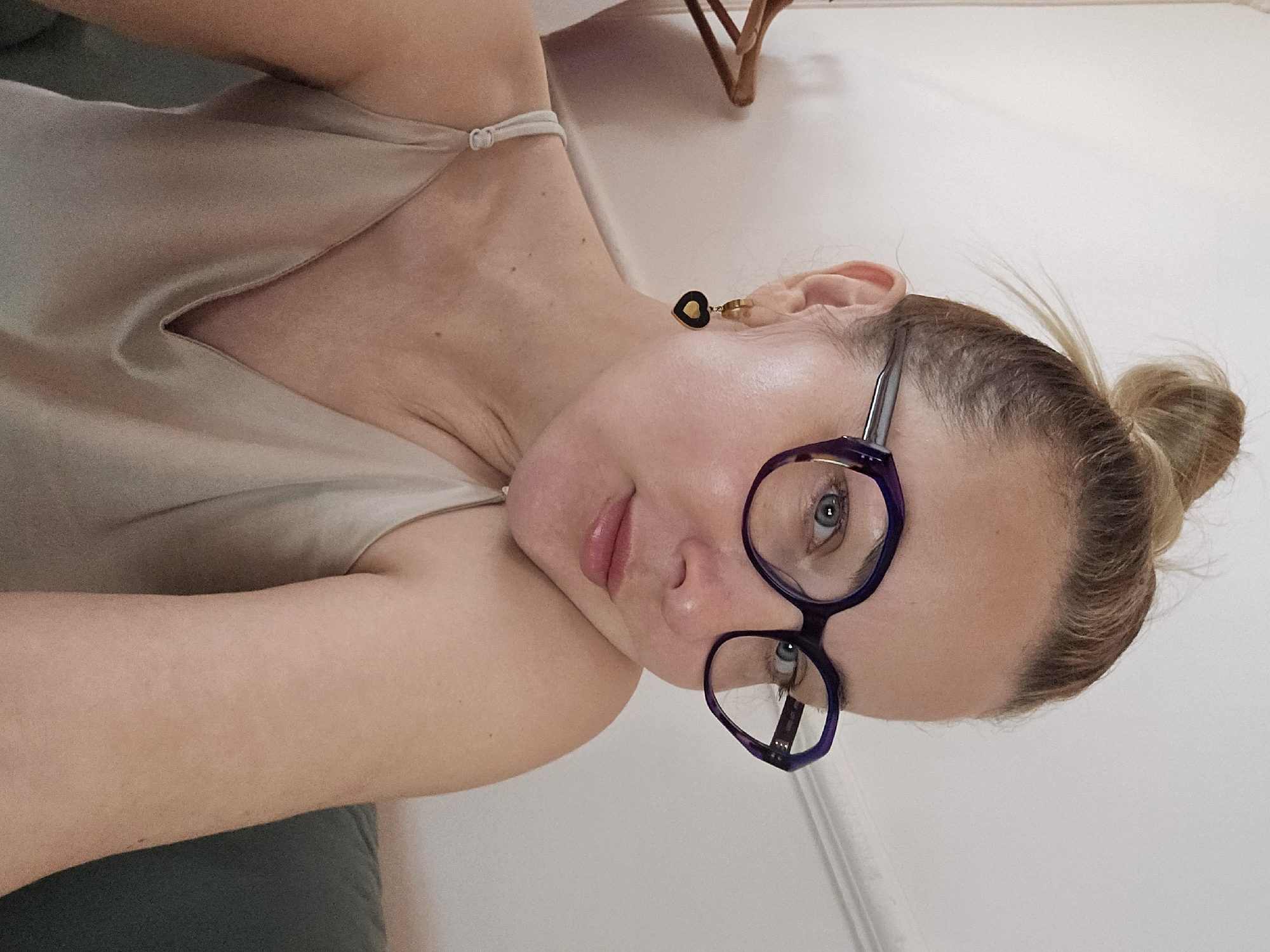 A woman with blonde hair pulled back into a bun is looking at the camera while wearing glasses with dark frames. She has light skin and is wearing a light-colored top. She has an earring on her left ear. The background is a white wall. The city of Sochi, Russia is not discernible from the image.