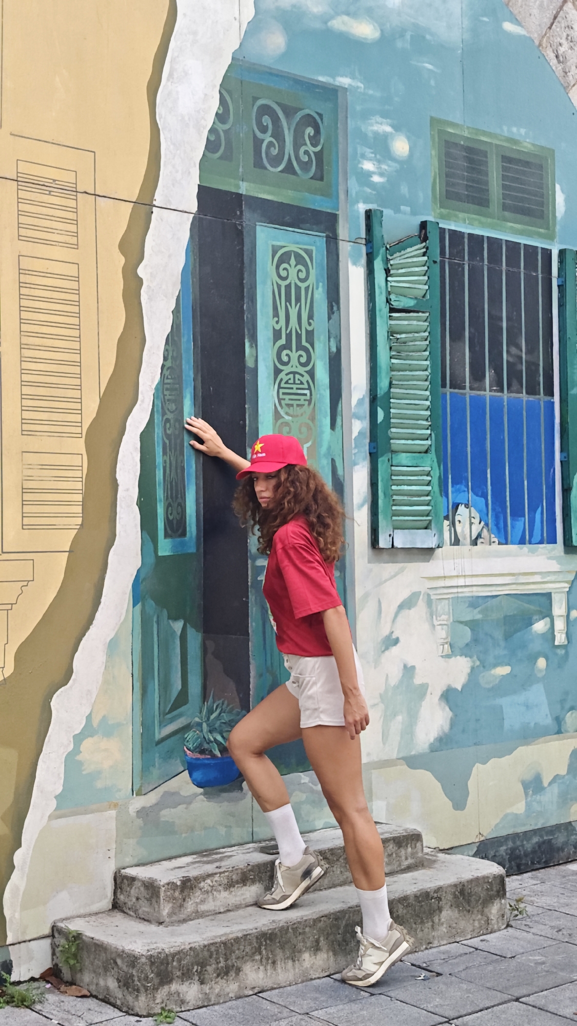 A woman in a red shirt, white shorts, a red baseball cap with a yellow star, white socks, and athletic shoes is climbing concrete steps next to a painted wall. The wall is painted with a torn-paper effect. On one side is a yellow building facade, and on the other, a blue-green rendering of a traditional Vietnamese building, complete with potted plants, windows with shutters, and a small child’s face painted inside one of the windows. The setting is urban, with a gray brick sidewalk at the base of the steps. The location is Hà Nội, Vietnam.