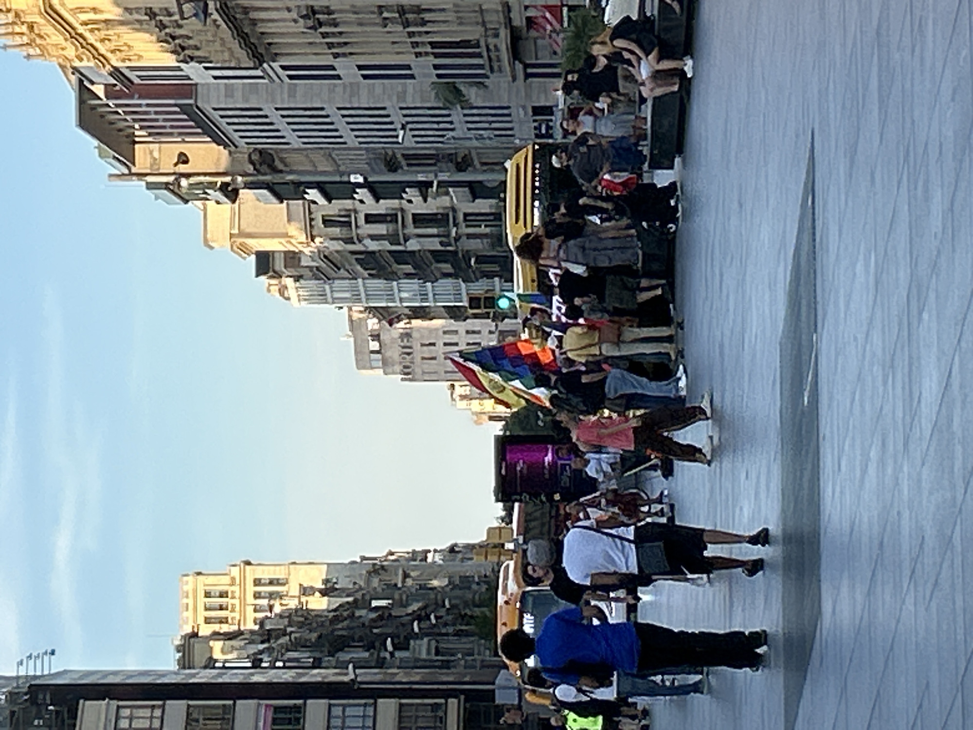 The image, taken in Barcelona, Spain, depicts an urban street scene on a clear, sunny day, likely during late afternoon or early evening given the golden light on the buildings. The perspective is from slightly above, looking down a wide paved area, possibly a pedestrian street or plaza.
In the foreground and midground, a group of people is gathered. Many are facing towards the right side of the frame, suggesting an event or procession. Several individuals are visible in various attire, some carrying flags that display multiple colors, including red, yellow, green, and blue. These flags suggest a demonstration or cultural event. One person near the center is seen holding what appears to be a musical instrument or equipment, possibly a violin or guitar, hinting at a performance.
To the left of the gathering, a yellow bus is partially visible, indicating active traffic or a stationary vehicle associated with the event. A green traffic light is also visible above the crowd, further placing the scene on a street.
The background is dominated by tall, multi-story buildings with numerous windows and architectural details typical of European cities. These buildings appear to be commercial or residential, lining both sides of the street. The sky above is clear and light blue. The ground is paved with large, light-colored tiles, possibly stone or concrete.
No specific recognizable landmarks of Barcelona are clearly visible, but the architectural style and urban density are consistent with the city. The overall atmosphere appears active and engaging, characteristic of a public gathering or demonstration. No specific text is clearly legible in the image.