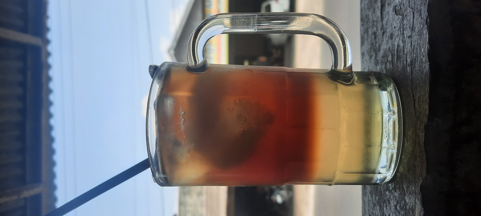 A close-up, vertical shot captures a tall glass mug filled with a layered iced beverage, placed on a dark, textured surface. A black straw emerges from the top of the drink, which has a lighter yellow layer at the bottom and a darker amber layer above, interspersed with ice cubes. The glass is slightly frosted from the cold. In the background, a blurred urban scene unfolds under a bright, partly cloudy sky. A corrugated roof structure is visible at the top of the image, and beyond that, a building and vehicles suggest a setting in Selopuro, Indonesia.