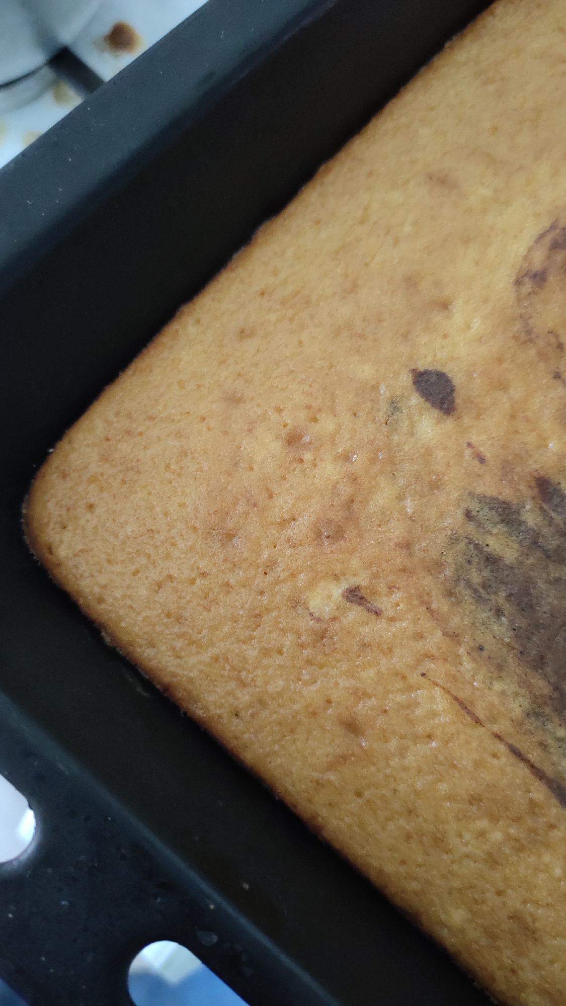 A close-up, high-angle shot shows a baked cake in a dark, rectangular baking pan. The cake has a golden-brown, slightly textured surface with some darker marbling patterns visible. The edges of the cake appear to be slightly browned. The baking pan is made of a dark material, possibly metal, with a matte finish. The background is out of focus but suggests a kitchen environment with hints of white and brown surfaces, possibly a stove or countertop. The lighting is even and natural, highlighting the texture of the cake.