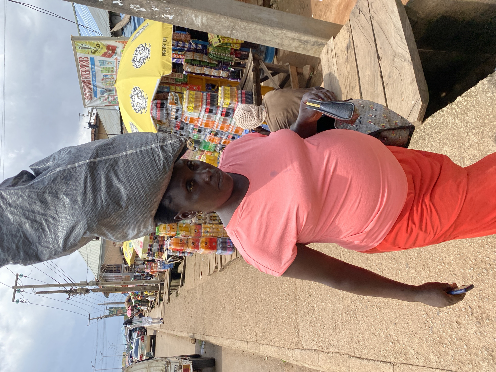 A pregnant Black woman walks along a sidewalk in Bukuru, Nigeria, carrying a large, gray, woven sack on her head. She wears a pink t-shirt and a long, orange skirt. Her left hand holds a dark-colored handbag, and her right hand holds a blue smartphone. The background features a bustling street market with stalls selling bottled drinks and other goods. A yellow umbrella with a tiger logo shades one of the stalls. In the distance, cars are visible on the road, and utility poles line the street. The sky is overcast.