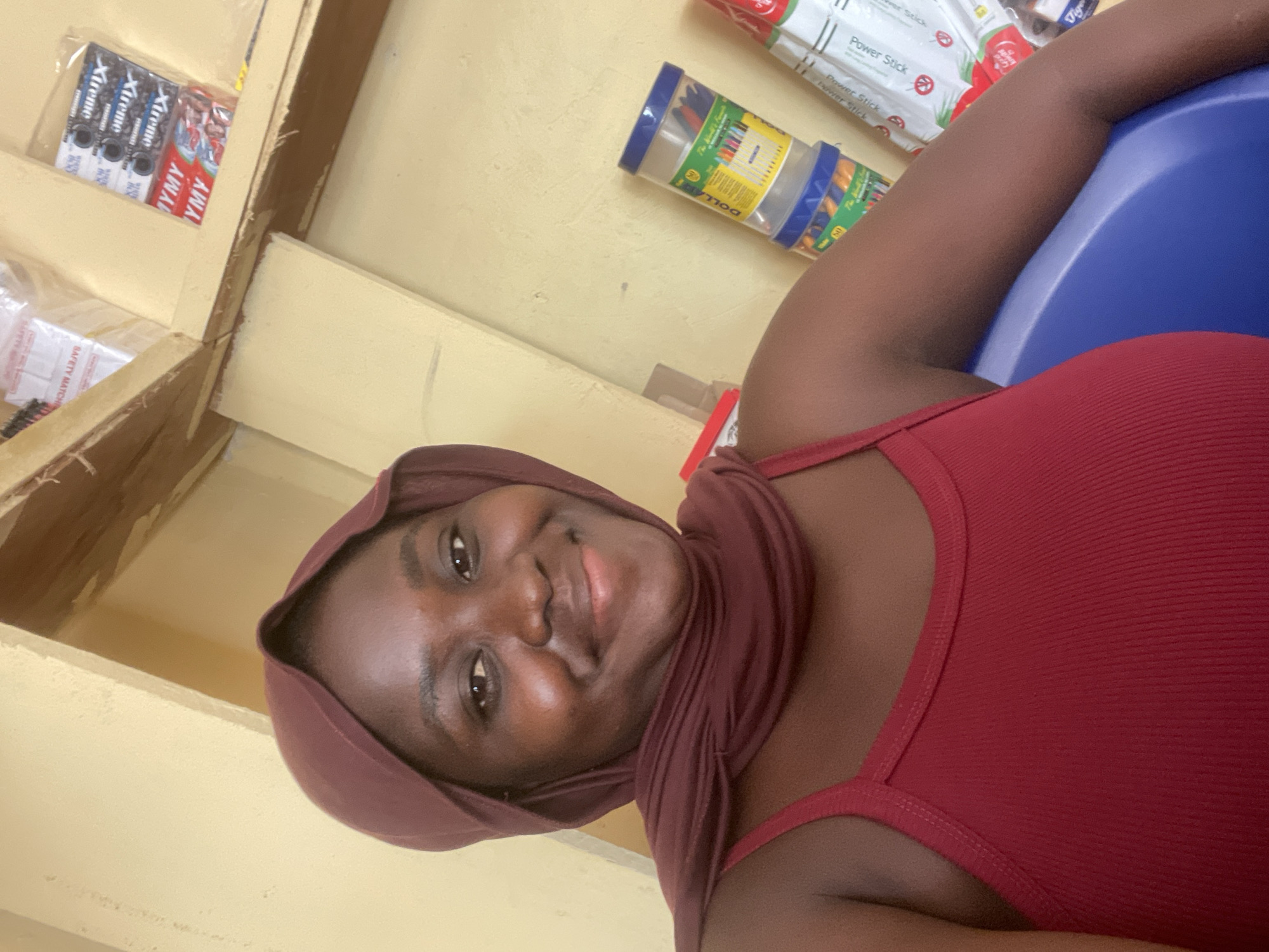 A dark-skinned woman wearing a maroon headscarf and a red tank top is pictured in a room with yellow walls and shelves. The shelves hold various items, including boxes of matches, packages of batteries labeled "Xtremic," and containers of pens or markers. The woman is smiling and looking towards the camera. She is seated on a blue surface, possibly a chair or stool. The environment appears to be a shop or a storage area.