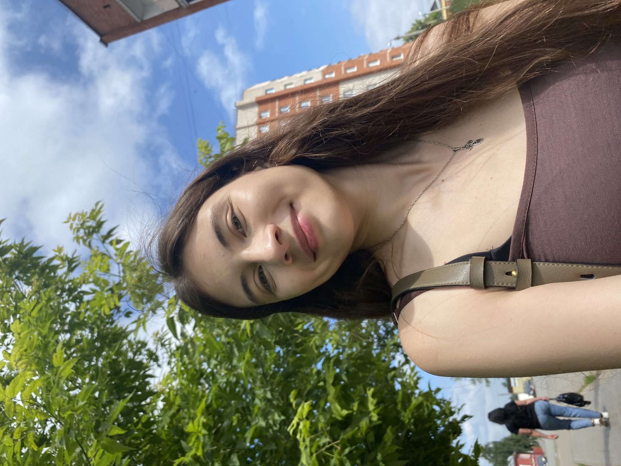 The image is a selfie taken outdoors in Votkinsk, Russia. A young woman with long brown hair and a brown tank top is in the foreground, smiling at the camera. She is wearing a silver necklace with a cross pendant. Behind her, a tree with green leaves is on the left, and a brick building is on the right. A person in a black shirt and blue jeans is walking away from the camera on a sidewalk in the background. The sky is blue with some clouds. The scene appears to be a residential area in Votkinsk.