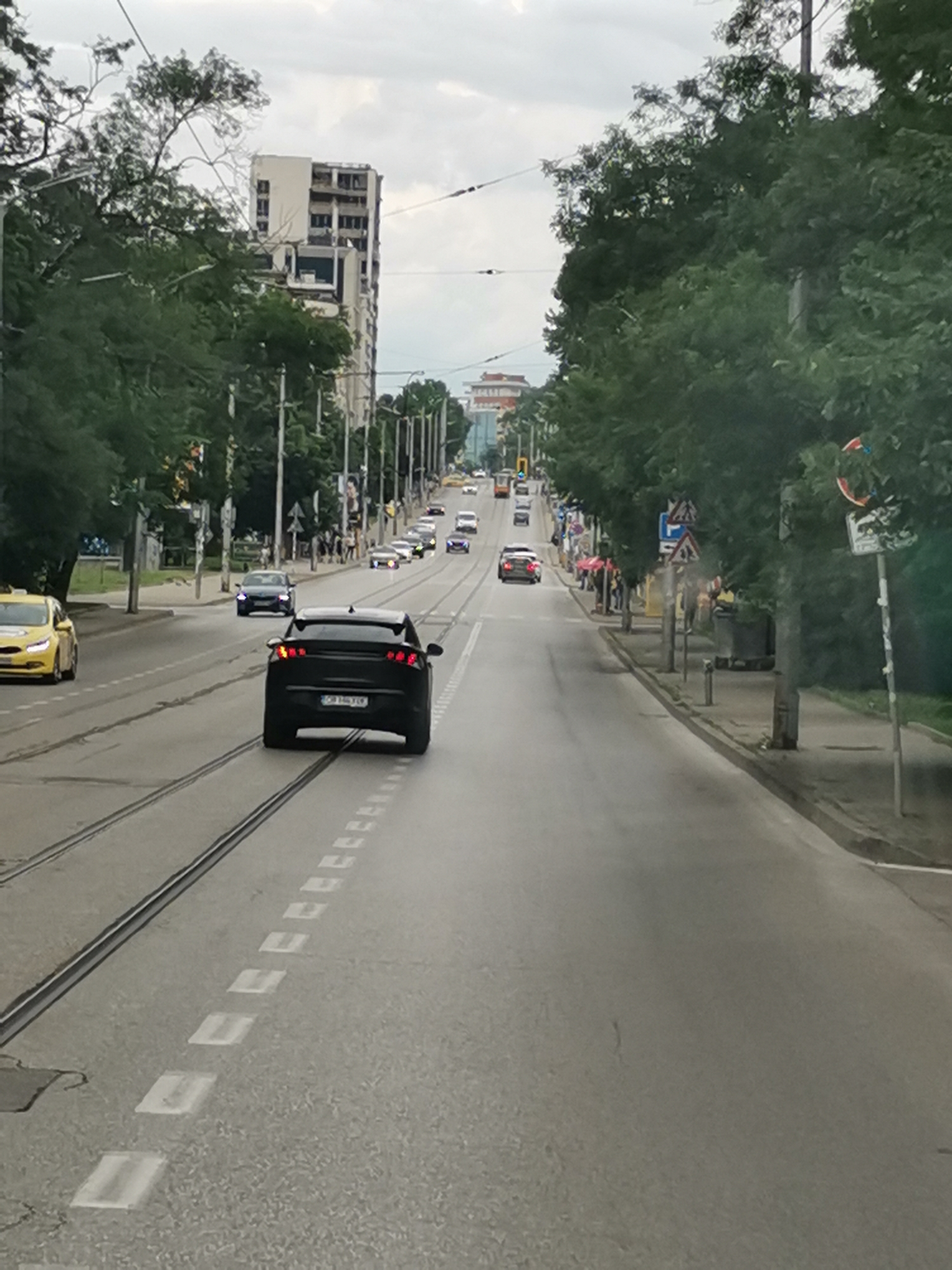 The image depicts a street scene in Sofia, Bulgaria. A black car is in the foreground, driving down the street. A yellow taxi is to the left of the black car, also moving forward. Several other cars are visible further down the road, as well as a tram. Trees line the street on both sides, and buildings are visible in the background. The sky is overcast. The scene appears to be a typical urban environment.