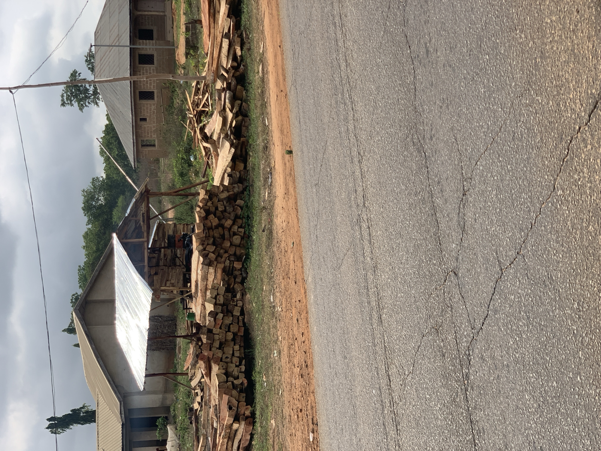 Along a cracked asphalt road in Kabba, Nigeria, a pile of roughly hewn timber and stacked wooden blocks rests against a dirt shoulder. A corrugated metal roof supported by wooden posts covers a section of the wood pile. Behind the structure are buildings with simple construction, appearing unfinished in places. Trees and overcast skies complete the backdrop. There are no people visible in the scene.