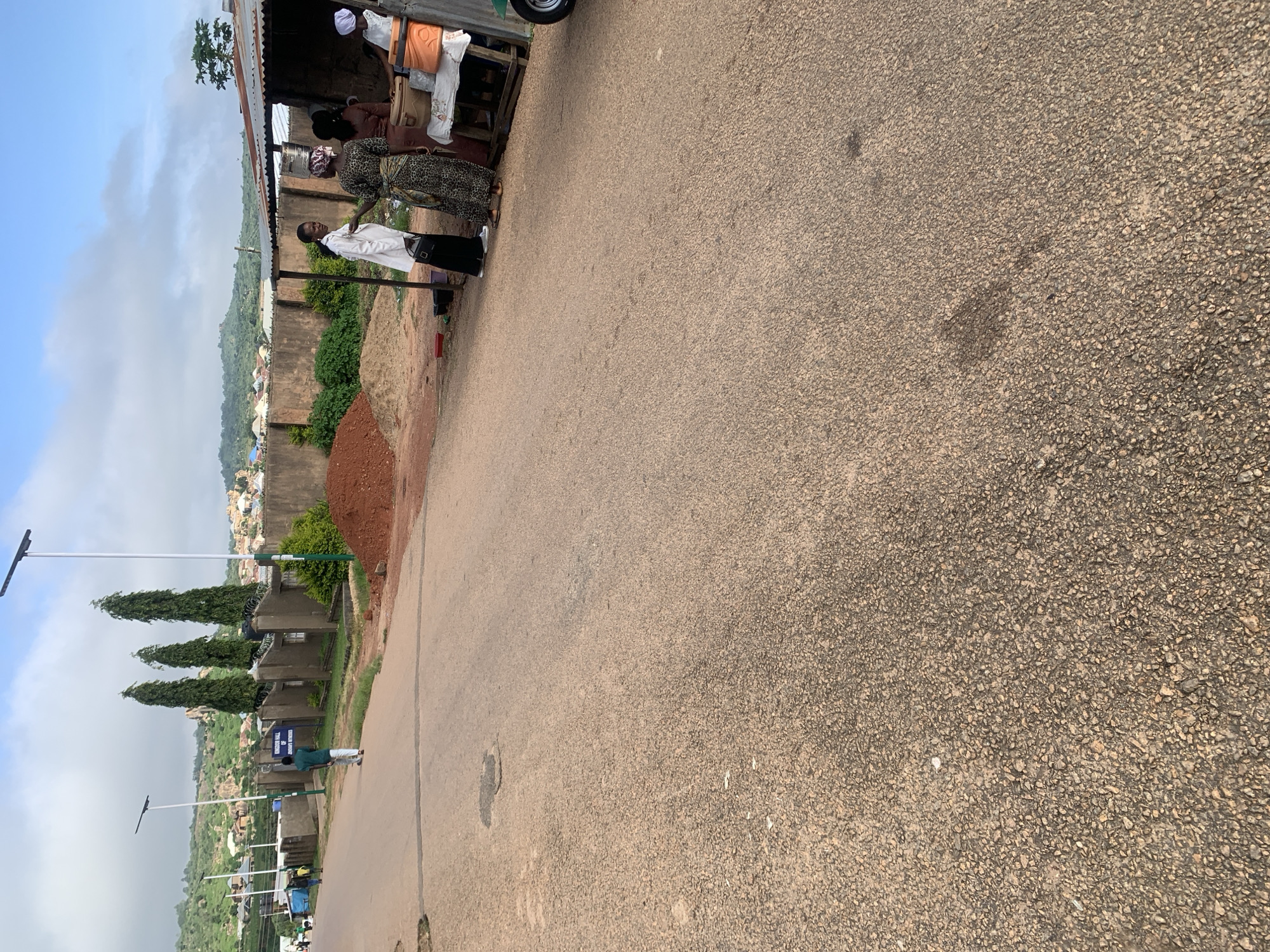 An outdoor, medium shot captures a paved road leading towards a cluster of buildings and a low wall, set against a backdrop of rolling hills under a bright sky with scattered clouds. On the right side of the road, a small, open-air market stall is set up under a corrugated metal roof. Three women are visible interacting at the stall. One woman, wearing a white coat and black pants, stands on the left, holding a black bag. Another woman, in a patterned dress, stands to her right, facing the first woman. A third woman, in a lighter colored dress and head covering, is partially visible behind them, as is a fourth woman with a white head covering, seated and facing away. Various containers and items are displayed on the table of the stall. A pile of reddish-brown soil or dirt is situated on the left side of the road, near a short, dark green bush. Further down the road on the left, three tall, slender evergreen trees stand behind a wall, with a blue sign attached to a pole in front of them. Two individuals are walking away from the camera on the left side of the road, one in a green shirt and light-colored pants, and the other in a yellow shirt and dark pants. The road surface is asphalt, showing signs of wear and a few dark patches. Solar-powered streetlights are visible along the left side of the road. The overall scene suggests a daytime setting in a semi-urban or rural environment.