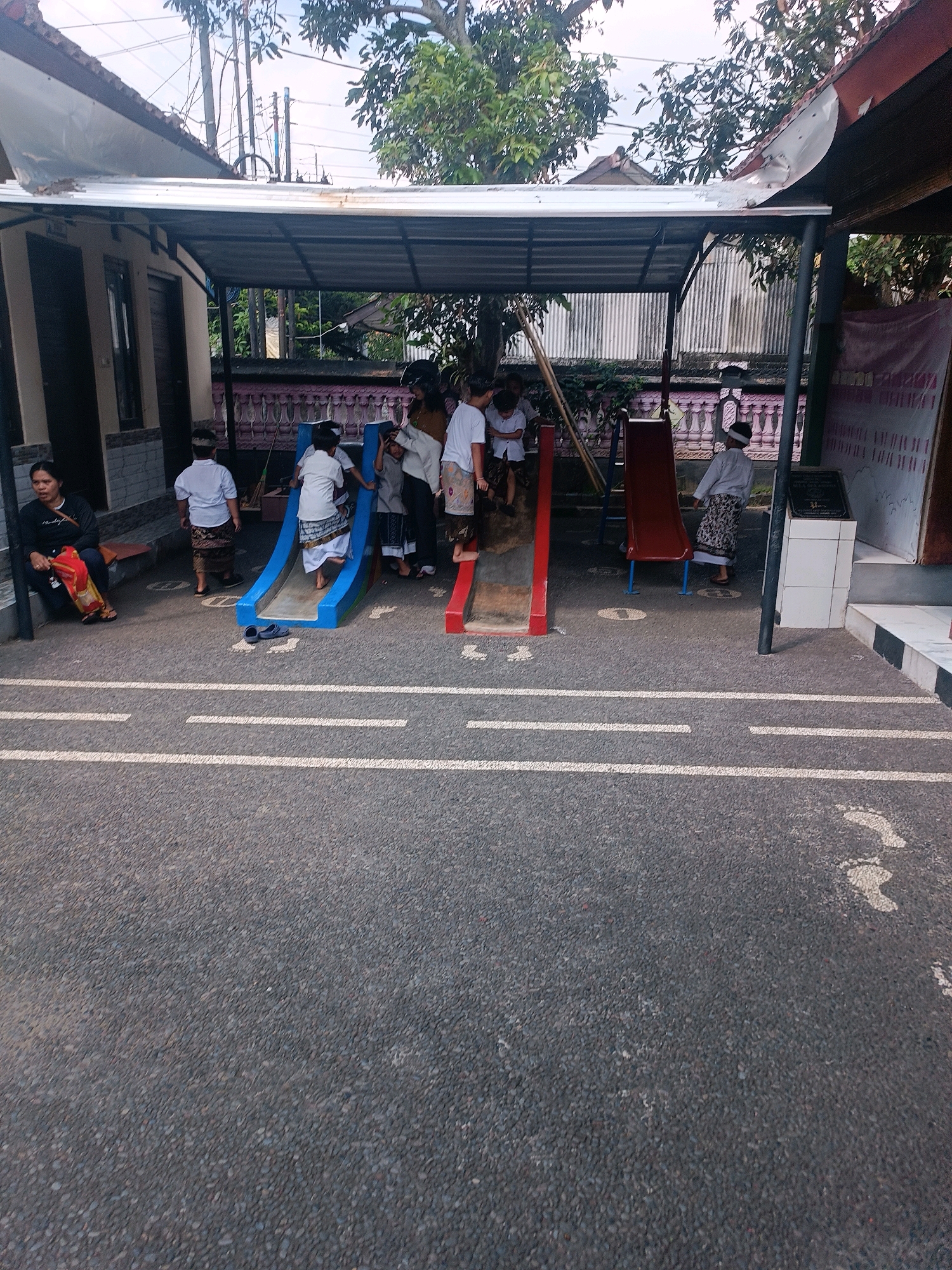 In Tabanan, Indonesia, an outdoor play area is captured. A woman sits on a low bench to the left, holding a red bag. In the center, a blue slide has children on it, with one child ascending and others at the top. A red slide is visible to the right, with a child sliding down. Several children, appearing to be school-aged, are dressed in white shirts and patterned sarongs. The ground is asphalt with painted white lines and footprint markings. A covered shelter with a metal roof extends over the play area. In the background, a building with a dark door, corrugated metal siding, trees, and power lines are visible. A banner hangs on the right side.