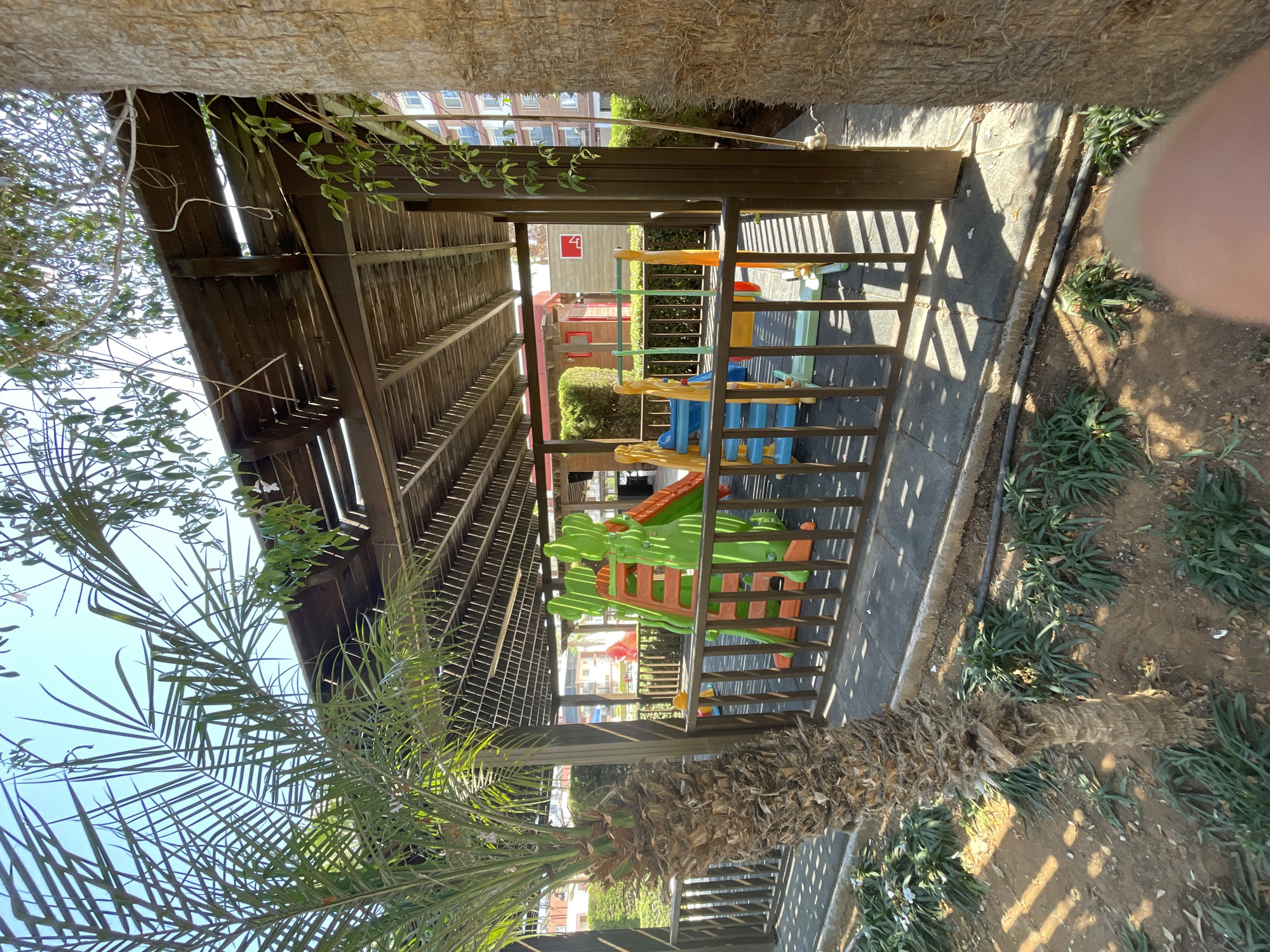 The image depicts a children's playground located in Trikomo, Northern Cyprus. The playground is situated under a wooden pergola, providing shade. It features brightly colored play structures, including slides and climbing elements, in green, orange, blue, and yellow. A brown wooden fence encloses the play area. To the left, a palm tree stands prominently, with its trunk and fronds visible. In the background, there are buildings and lush green foliage. The ground surface of the playground appears to be paved with dark tiles. The scene is bathed in natural sunlight, casting shadows from the pergola and trees.
