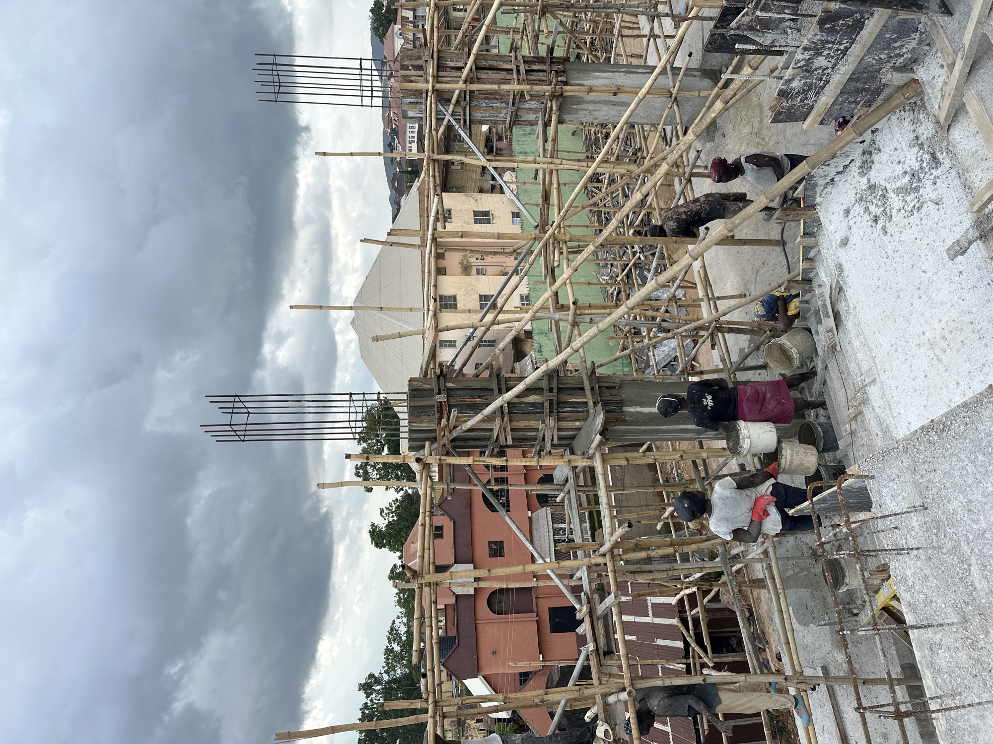 The media file depicts a construction site with several people working. The scene is dominated by scaffolding made of bamboo and wood, supporting concrete structures and rebar. In the foreground, two individuals are visible, one wearing a white shirt and red gloves, appearing to be engaged in some form of manual labor. Another person is seen further down, also working. Various buckets and construction materials are scattered around the site.
In the background, several buildings can be seen, suggesting an urban or semi-urban environment. The sky is overcast with dark clouds, indicating potentially inclement weather, possibly late afternoon or evening. The overall atmosphere of the scene is one of active construction work. There is no visible text in the image.