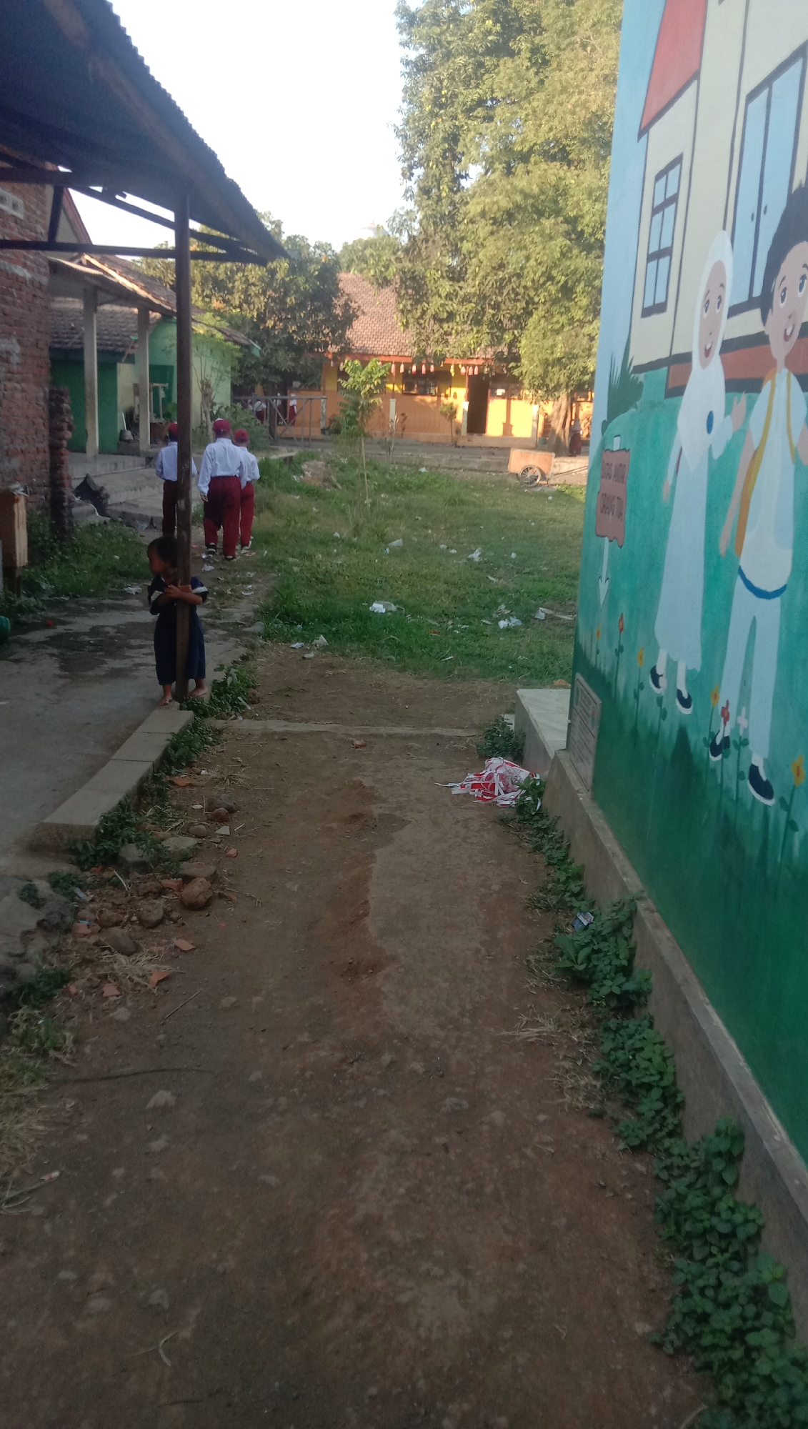 The image depicts an outdoor scene in Karanganyar, Indonesia, likely a schoolyard. In the foreground on the left, a young child in a dark blue outfit stands near a wooden pole, looking towards the left. Further back on a dirt path, three school-aged children in white shirts and red pants walk away from the viewer.
The environment includes a building with a corrugated metal roof on the far left. Behind the children, there is a grassy area with some scattered trash. In the background, more buildings with tiled roofs are visible, surrounded by dense green trees. To the right of the frame, a colorful mural of two children, one in a hijab and the other in a yellow shirt, is painted on a wall. The ground is a mix of dirt and some paved areas. The lighting suggests it is either early morning or late afternoon, with sunlight casting shadows.