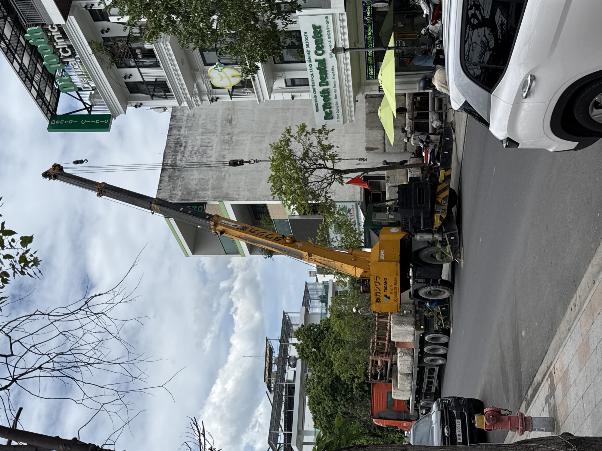 The image shows a construction scene in Nha Trang, Vietnam, specifically on a street in front of a dental clinic. A yellow Tadano crane is actively lifting concrete blocks from a flatbed truck. Several workers are present, some wearing hard hats, observing the operation or engaged in other tasks. The dental clinic, named "Dr. Tooth Dental Center," has signage in Vietnamese and English. The street is paved, with a sidewalk and a fire hydrant visible. Other vehicles, including a black Ford SUV and motorcycles, are parked or driving on the street, indicating a normal day in the city despite the construction work. Trees and buildings line the street, contributing to the urban environment. The sky is partly cloudy.