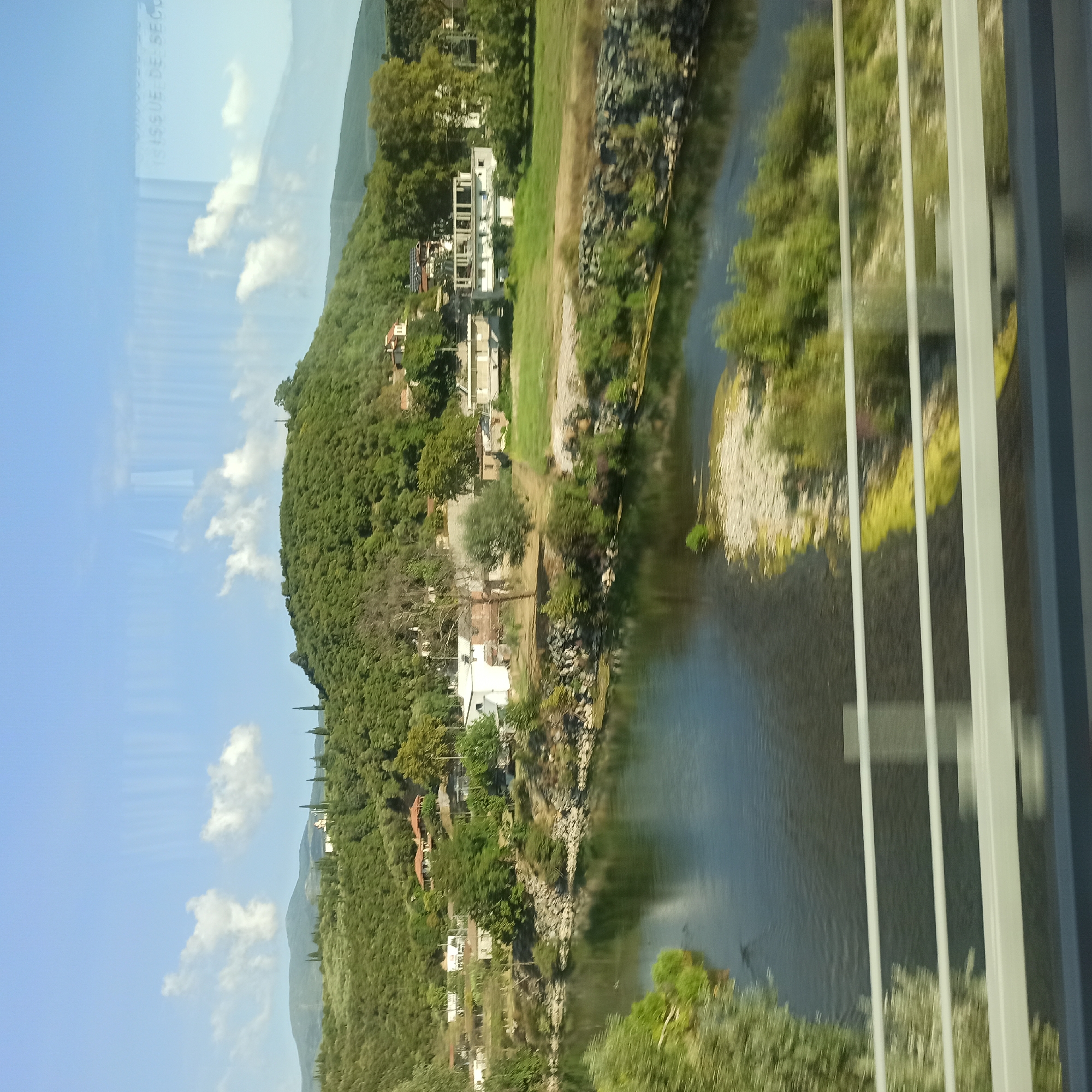 The image captures a scenic view of a river flowing through a rural landscape in the Municipal Unit of Kato Olympos, Greece. A lush green hill, covered in trees and dotted with a few houses, rises behind the riverbank. The river itself is dark and reflects the sky and surrounding greenery. Along the riverbank, there are rocks and vegetation. In the foreground, there are white railings, suggesting the image was taken from a bridge or overpass. The sky is blue with a few scattered white clouds. The overall impression is one of tranquility and natural beauty.