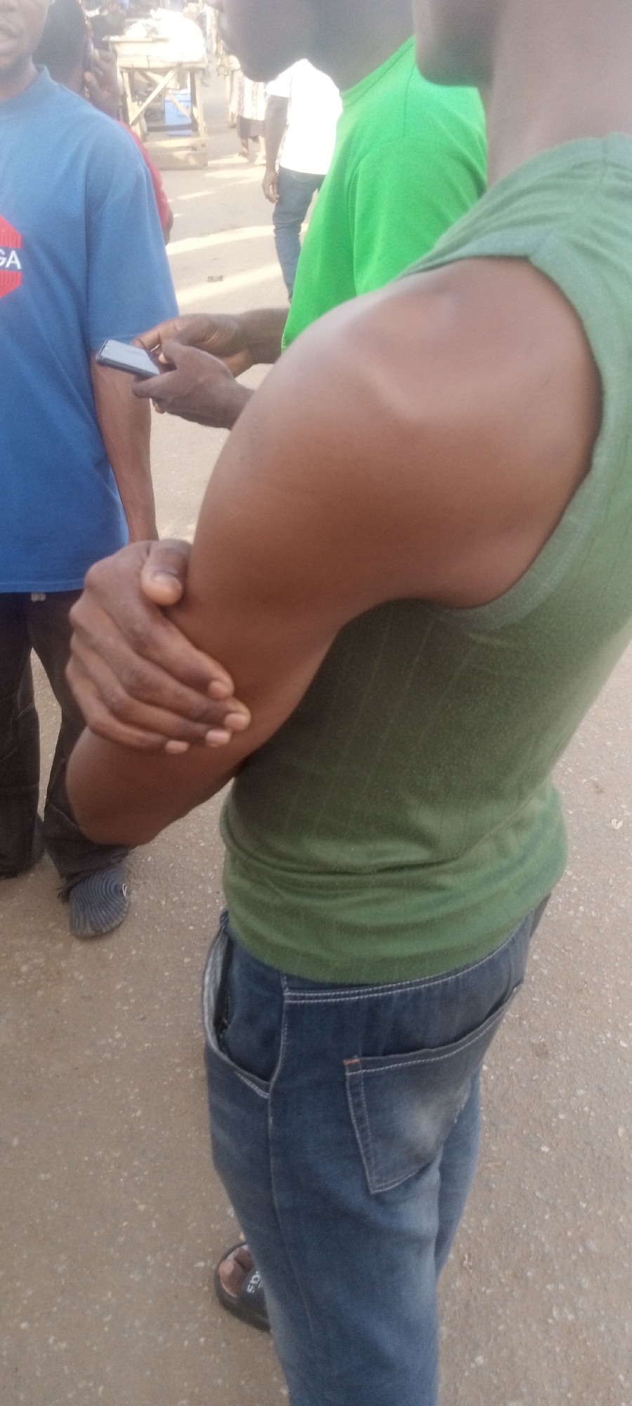 In Narayi, Nigeria, a medium-angle outdoor shot depicts several individuals in a public setting. The focal point is a muscular man wearing a green tank top and blue jeans with sandals. His left arm is flexed, and he's holding his forearm. To his left is a man in a blue t-shirt holding a smartphone, likely taking a photo. Another person wearing a green shirt stands next to the main subject. The background includes other blurred individuals and what appears to be street furniture or vendor stalls. The ground is light brown, suggesting a paved or dirt surface.