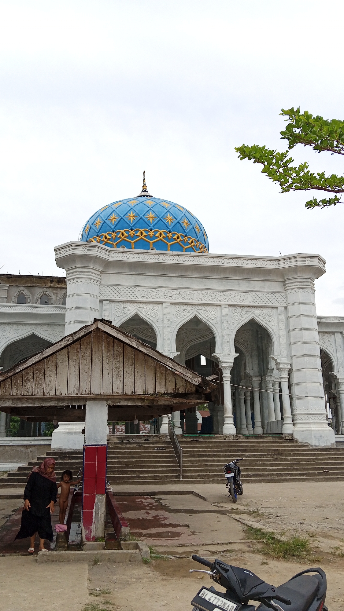 The image depicts a mosque in Beunyot, Indonesia, with a prominent blue and gold dome. In the foreground, a woman in a black hijab and a child stand near a water source with tiled pillars. Several stone steps lead up to the mosque's entrance, which features arched doorways and white columns. A wooden structure with a dilapidated roof is positioned in front of the steps. Two motorcycles are visible, one parked near the steps and another partially visible in the lower right corner. The sky is overcast. The overall scene suggests a public area adjacent to a religious building.