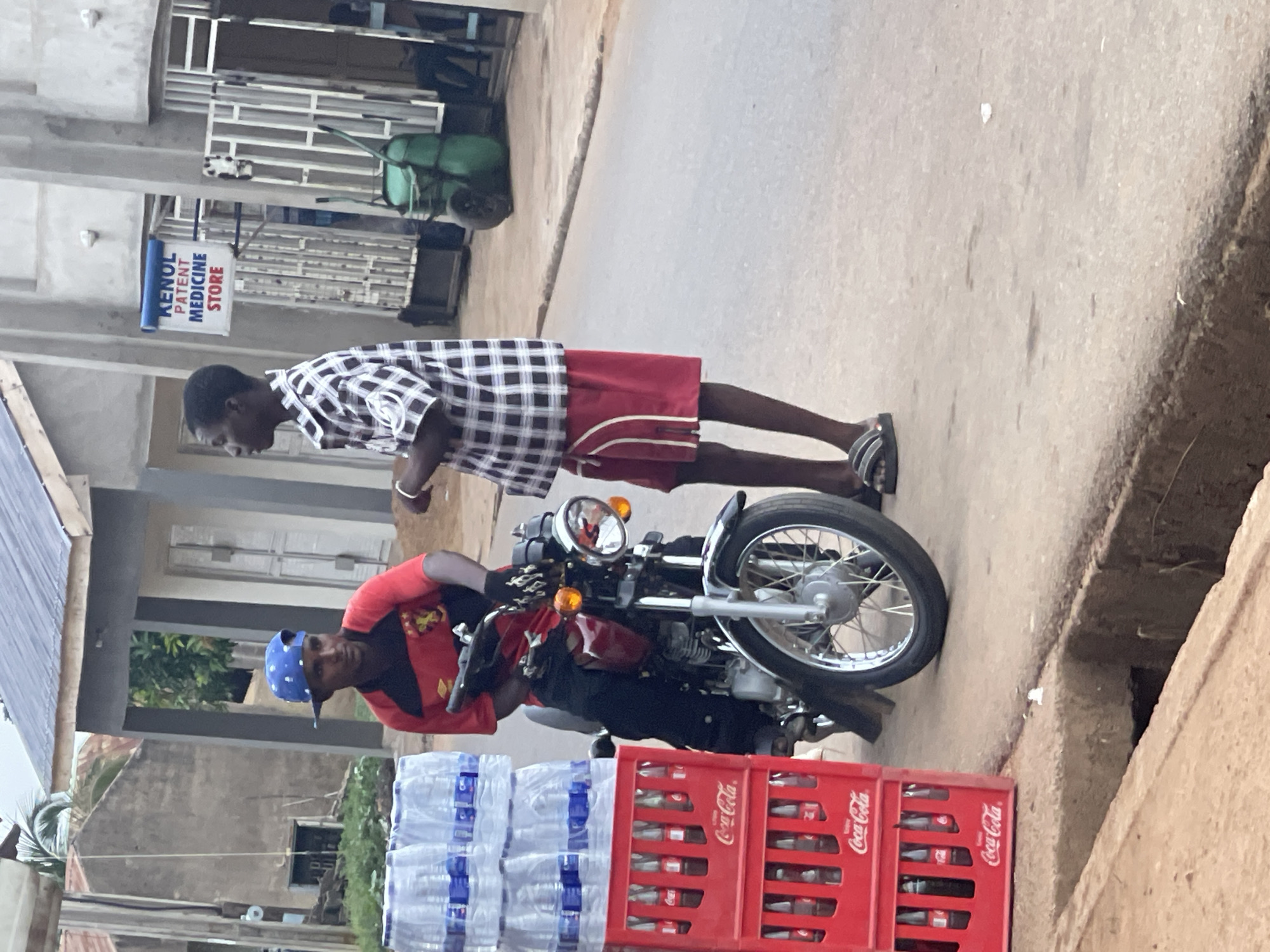 A man in a red shirt and blue hat is seated on a red motorcycle, positioned at the edge of a paved street in Akwanga, Nigeria. A stack of water bottles and red Coca-Cola crates are located beside the motorcycle. Another man, wearing a checkered shirt and red shorts, stands next to the motorcycle. A sign reading "KENOL PATENT MEDICINE STORE" is visible on a building in the background.