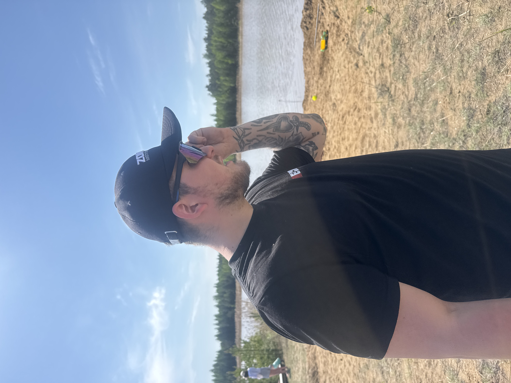 A person wearing a black t-shirt, black baseball cap, and sunglasses is standing on a sandy beach next to a body of water. The person is looking to the side, with their arm raised as if adjusting their glasses. In the background, there is a line of trees and blue sky with wispy clouds. There is some fishing tackle on the sand nearby. A person on a bicycle is visible in the background. This location is outside of Novyi, Russia, possibly on a bank of a lake.