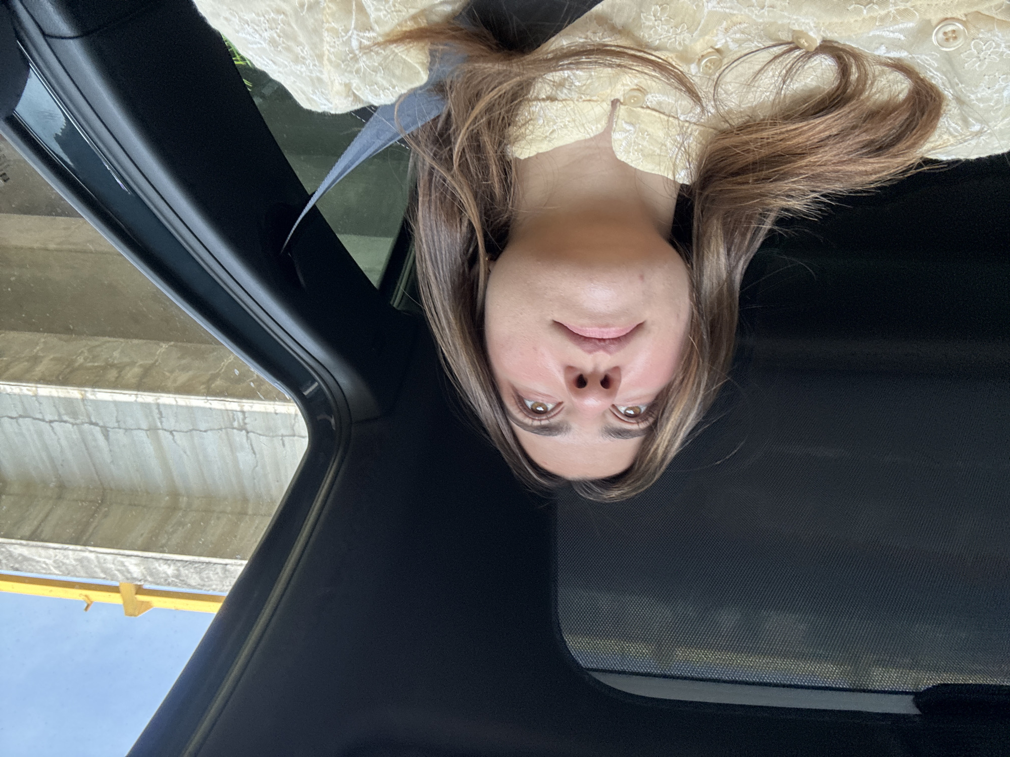 A woman is looking down at the camera from the sunroof of a car, seemingly at an upward angle. She has brown hair and is wearing a light-colored, patterned top. Outside the car, a structure with yellow elements is visible against a pale sky. The location is in the city of San Andrés Cholula, Mexico.