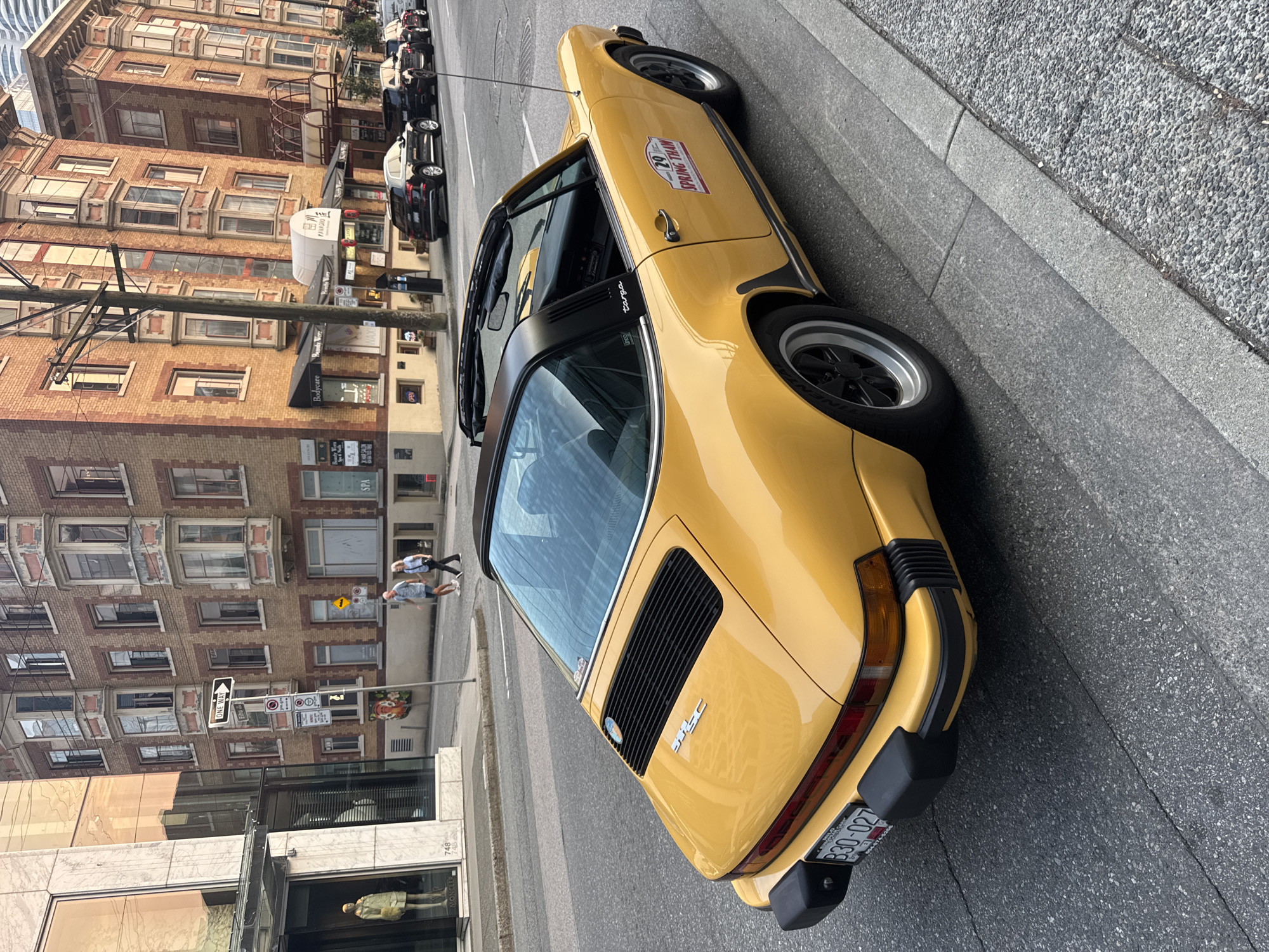 A yellow Porsche 911 Targa is parked on the side of a street in Vancouver, Canada. The car is viewed from the rear-left quarter. The car has a black targa top. A sticker on the passenger door reads "29 SPRING THAW". The license plate reads "B30-027". In the background, there are brick buildings with bay windows and a modern glass-fronted building. Pedestrians are walking on the sidewalk in front of the brick buildings. There are also several black cars parked along the street. The scene appears to be an urban environment.