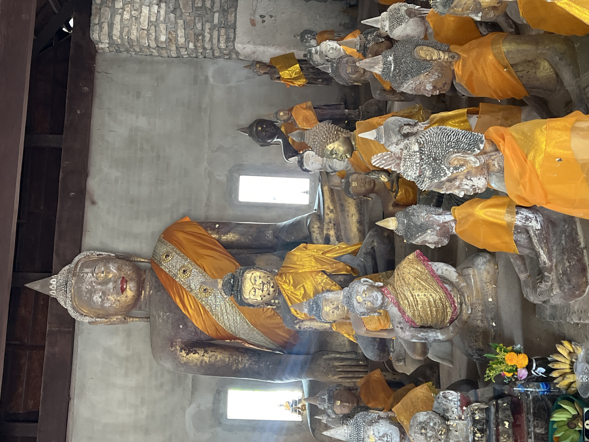 The image captures a multitude of Buddha statues at Wat Yai Chaimongkol, a Buddhist temple in Baan Harn, Thailand. The statues are predominantly in a reclining or seated position, draped in orange and gold cloths. The figures are weathered and appear aged.
The scene is indoors, within a structure with brick and concrete walls. Light enters through a window. At the base of the statues, there are offerings, including flowers and bananas. A variety of smaller religious objects are present. The wooden support structure of the building is visible.