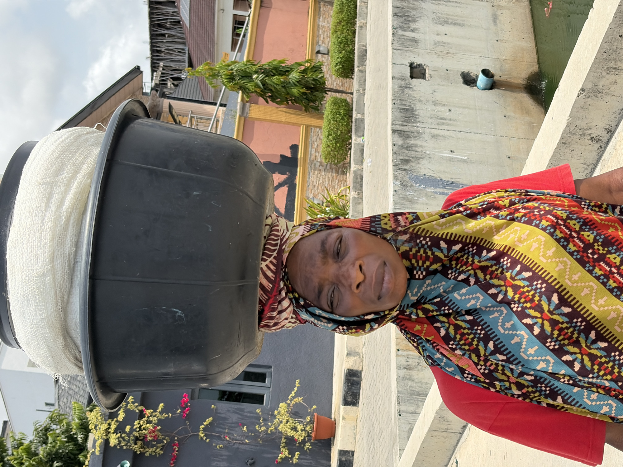 A woman is standing on what appears to be a city bridge in Igboefon, Nigeria. She is wearing a colorful headscarf and a red top. On her head, she is carrying a large black tub filled with a white, bundled material. A concrete bridge railing is visible to her right, with a waterway below. Behind her are buildings, greenery, and a section of an orange-colored structure. The sky is overcast.