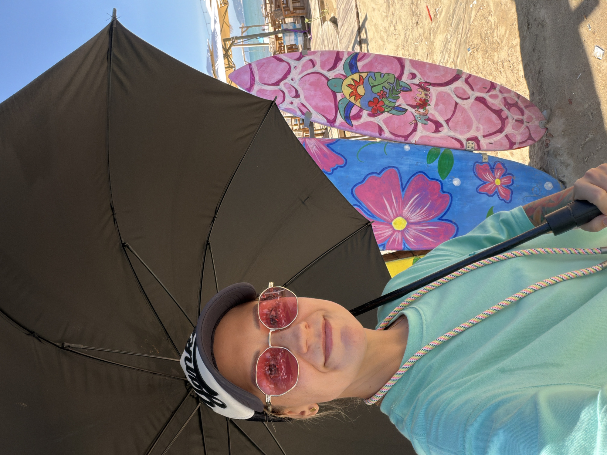 In Quy Nhon Ward, Vietnam, on a sunny day, a woman wearing a cap and pink sunglasses holds a black umbrella to shade herself. She is dressed in a mint green t-shirt and has a colorful cord around her neck. Two surfboards, one blue with pink flowers and another pink with a turtle design, stand behind her on the sandy beach. In the background, beach umbrellas, tables, and chairs are visible, along with the ocean and clear blue sky.