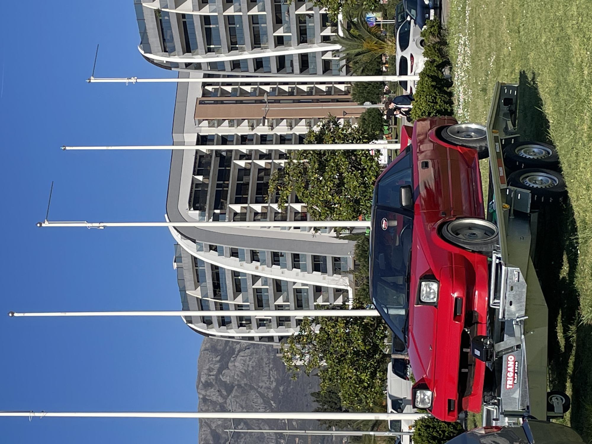 A red sports car is secured to a trailer in Bar, Montenegro. The trailer's tongue is visible, showing the brand TRIGANO. The surrounding area has a grassy lawn, a tree, and other parked vehicles. In the background, there's a modern, curved building and a distant mountain. Several light poles line the foreground under a blue sky.
