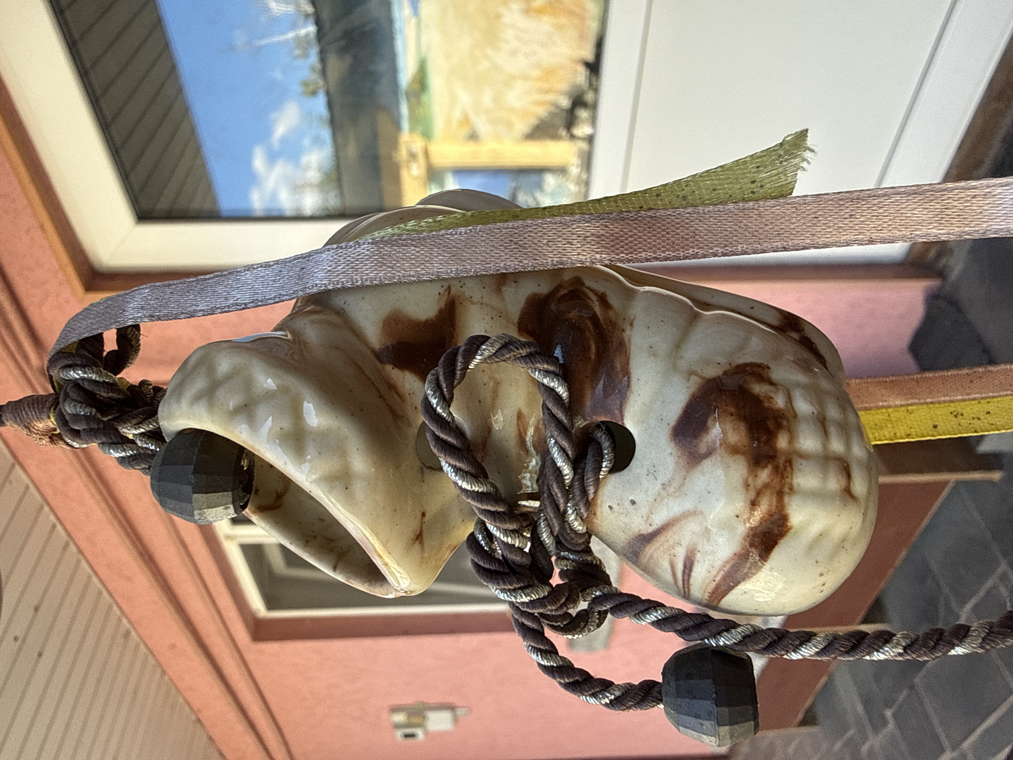 A close-up, low-angle shot focuses on a ceramic wind chime hanging outdoors. The wind chime is beige with brown swirls and has a carved, faceted black bead attached to a thick, dark brown rope with silver threads. The rope loops through holes in the ceramic. A thin, pink ribbon and a green ribbon with frayed edges are also attached to the wind chime. In the background, a pink wall with a window is visible, reflecting a blue sky with clouds. A white window frame and a part of a door are also in the frame. The ground beneath appears to be paved. The image seems to have been taken on a sunny day. This scene is located in Okhtyrka, Ukraine.