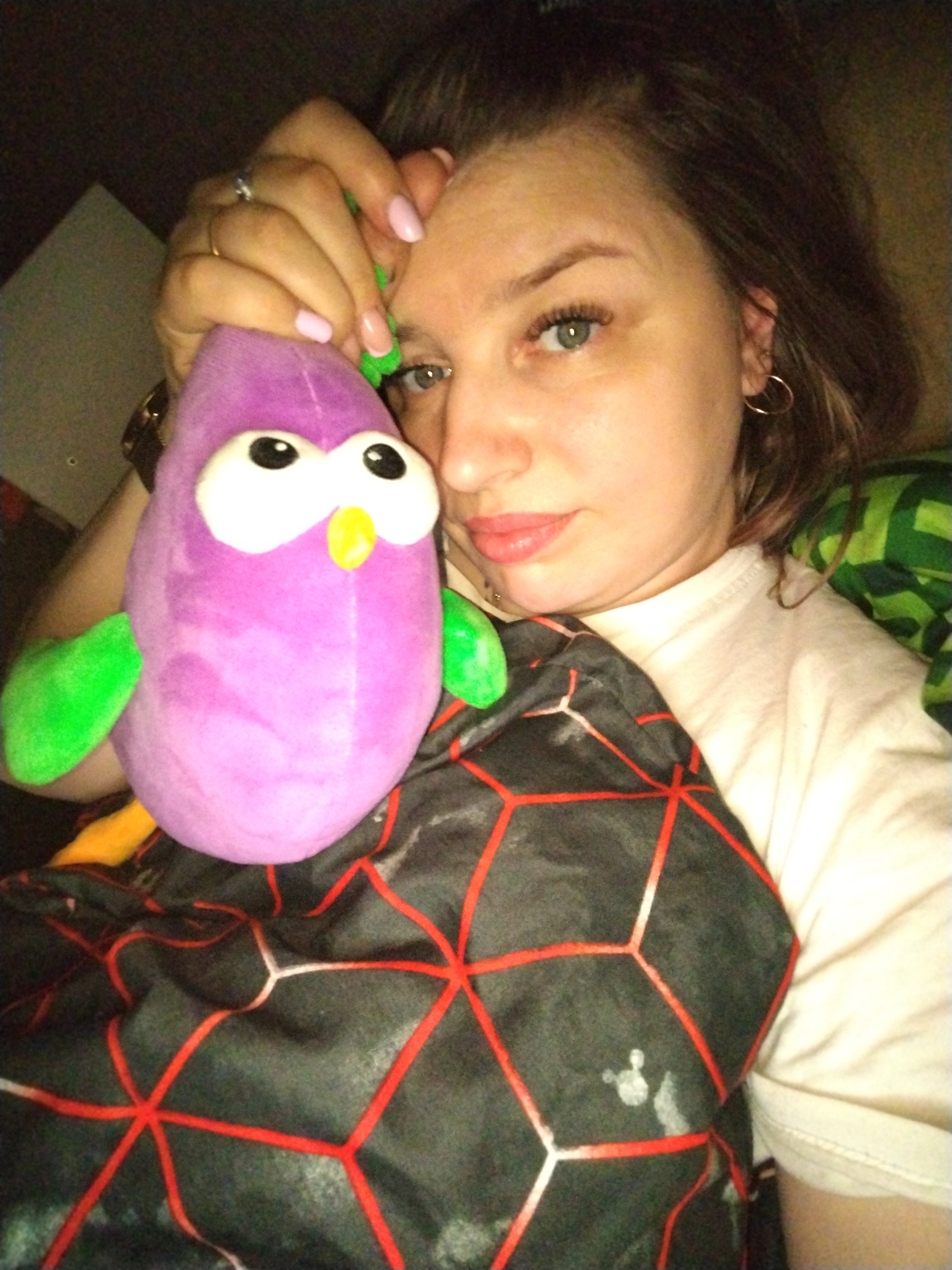 A young woman is depicted indoors, reclining and holding a purple plush toy. She is positioned in the foreground, looking directly at the camera with blue-green eyes. Her medium-length brown hair frames her face, and she has light pink nail polish on her manicured nails. A ring is visible on her left ring finger, and she wears a gold hoop earring in her right ear. She has a calm expression and is wearing a light-colored t-shirt.
The prominent plush toy is purple, shaped like a pear or bird, with large white eyes, black pupils, a small yellow beak, and green wings. The woman's left hand cradles this toy.
She appears to be partially covered by a black and red geometrically patterned blanket or fabric, featuring a subtle Mickey Mouse head silhouette. To her right, a glimpse of green, possibly checkered, fabric is visible. The scene is dimly lit, suggesting an indoor setting, potentially during the evening or night. The overall mood is relaxed and casual. No visible text is present in the image.