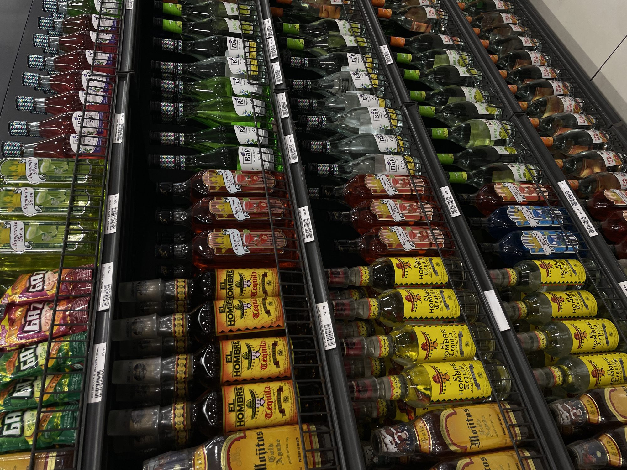This is a mid-angle, eye-level shot of a retail store aisle, specifically within a supermarket in Davao City, Philippines. The shelves are stocked with a variety of alcoholic beverages. The primary subjects are bottles of tequila, mojitos, and various flavored liqueurs and spirits, predominantly in glass bottles of various colors including clear, green, yellow, and red.
The products are arranged neatly on black metal shelves. The lighting appears to be artificial, typical of a supermarket interior, with no direct sunlight visible. The background is out of focus, but suggests additional retail shelving and possibly other sections of the store. There are no people clearly visible in the frame, and no products are being handled or interacted with. The overall impression is a well-stocked retail display of alcoholic drinks.
