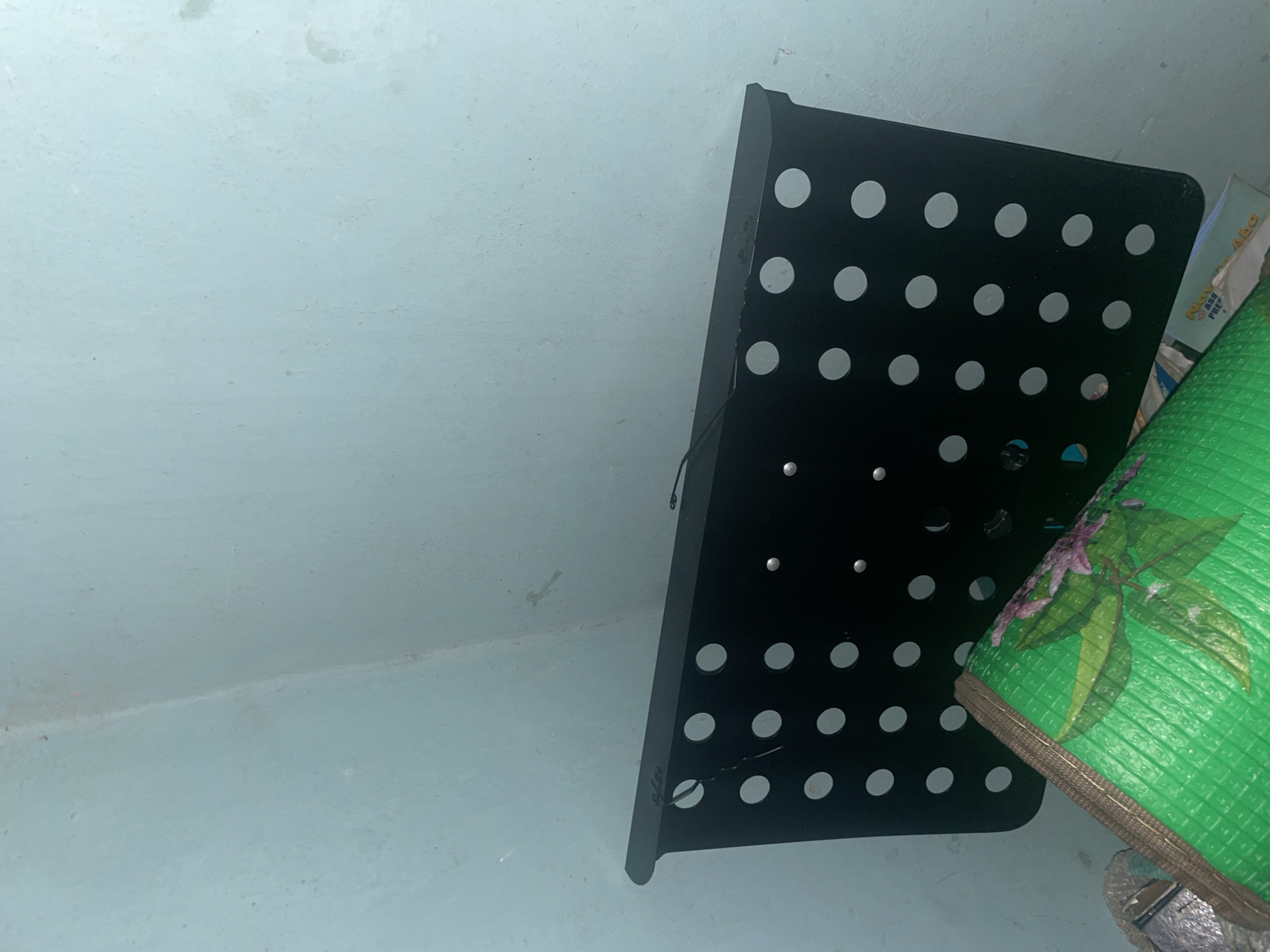 A black perforated metal stand is positioned vertically in a corner against a light blue wall. The stand has a series of evenly spaced circular holes across its surface. In the foreground, a rolled-up green mat with a floral pattern is visible. The location is identified as Jalingo, Nigeria. The environment appears to be indoors, possibly a storage area or a utility space, given the simple wall and the presence of stored items.